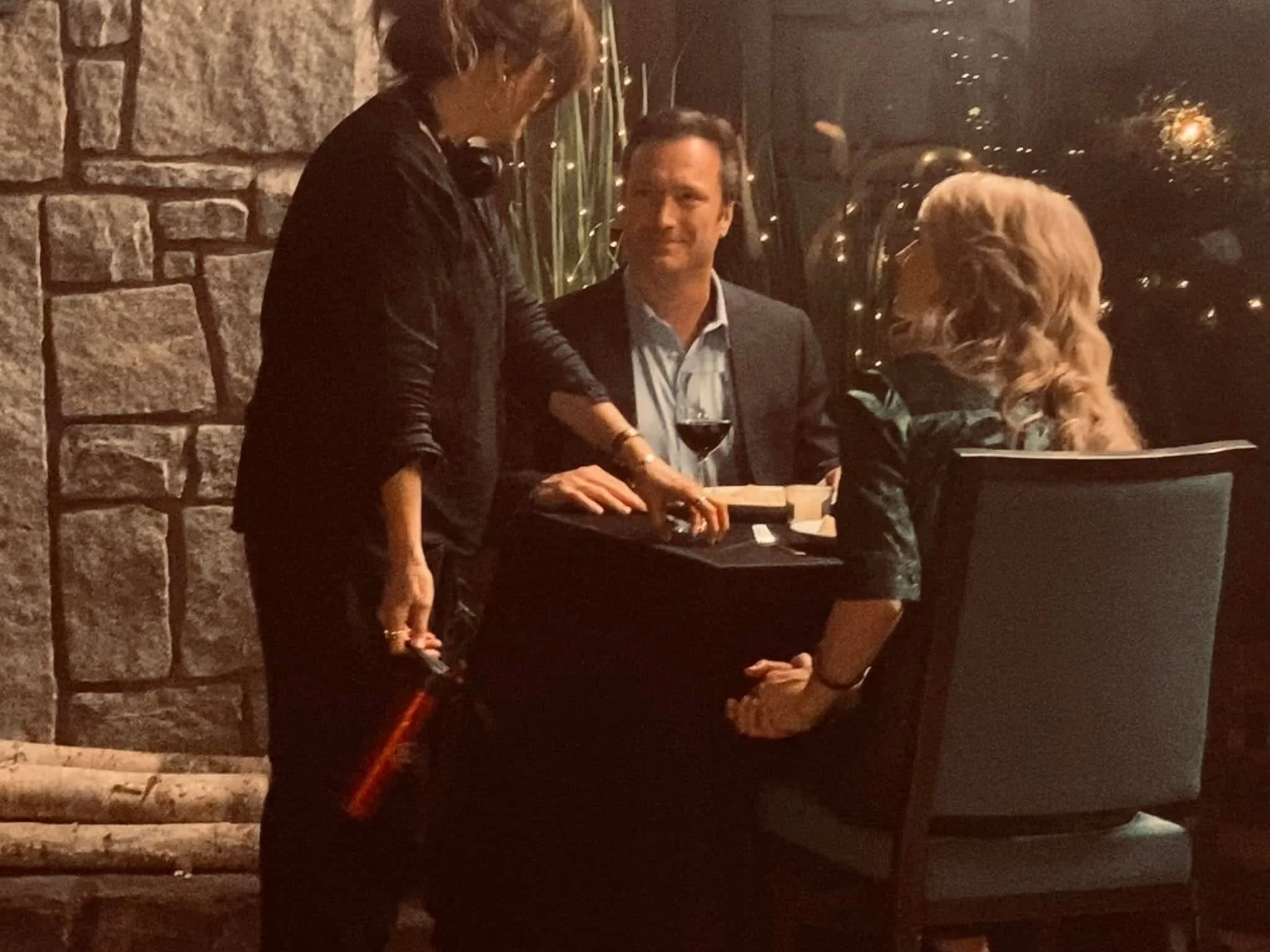 Three people are sitting at a table in a dimly lit restaurant or bar, with a woman standing next to the table. A man in a suit is holding a glass of red wine, and a woman with long, curly hair is sitting across from him, partially visible. The backgr