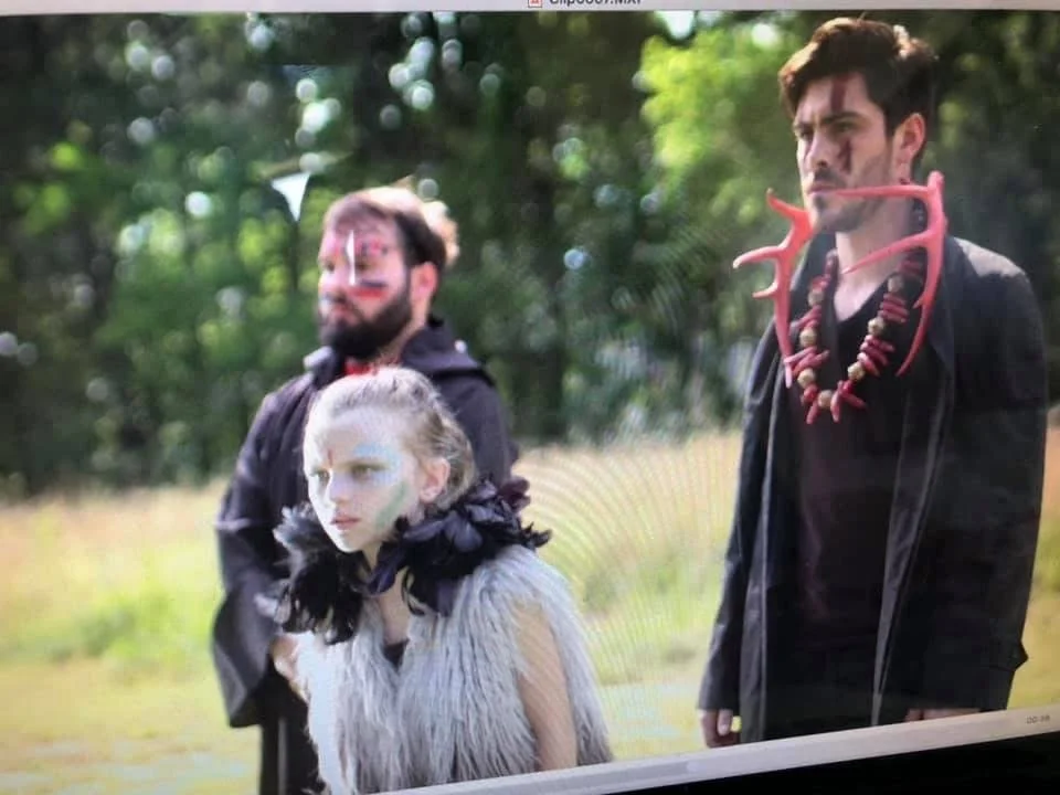 Three people outdoors dressed in dark clothing with face and body paint, and wearing nature-inspired accessories like a red antler necklace and black feathered cape, in a wooded area.