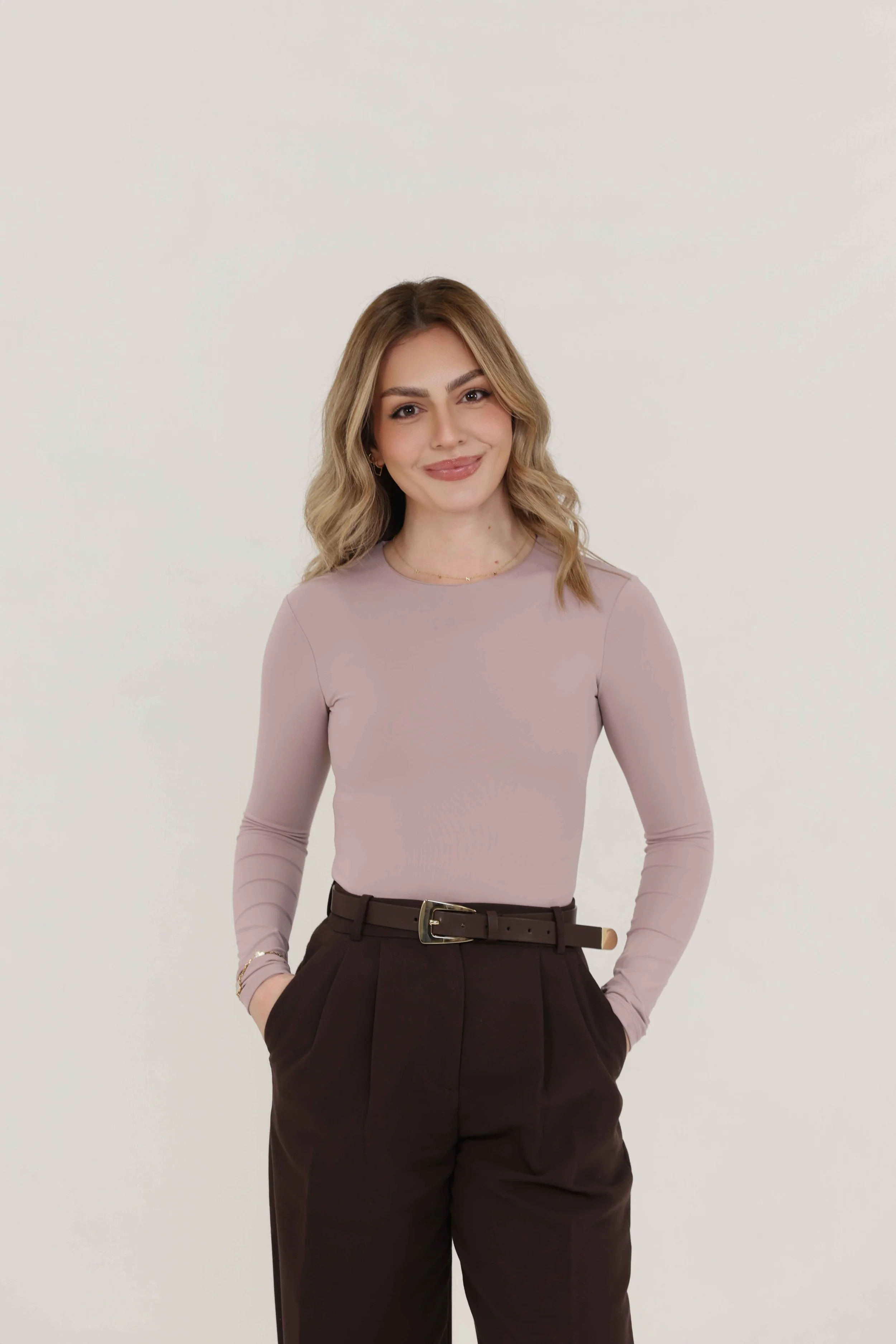 A young woman with wavy blonde hair and a warm smile, wearing a light purple long-sleeve top, black high-waisted trousers with a belt, and accessorized with small earrings and a delicate necklace, stands against a plain white background.