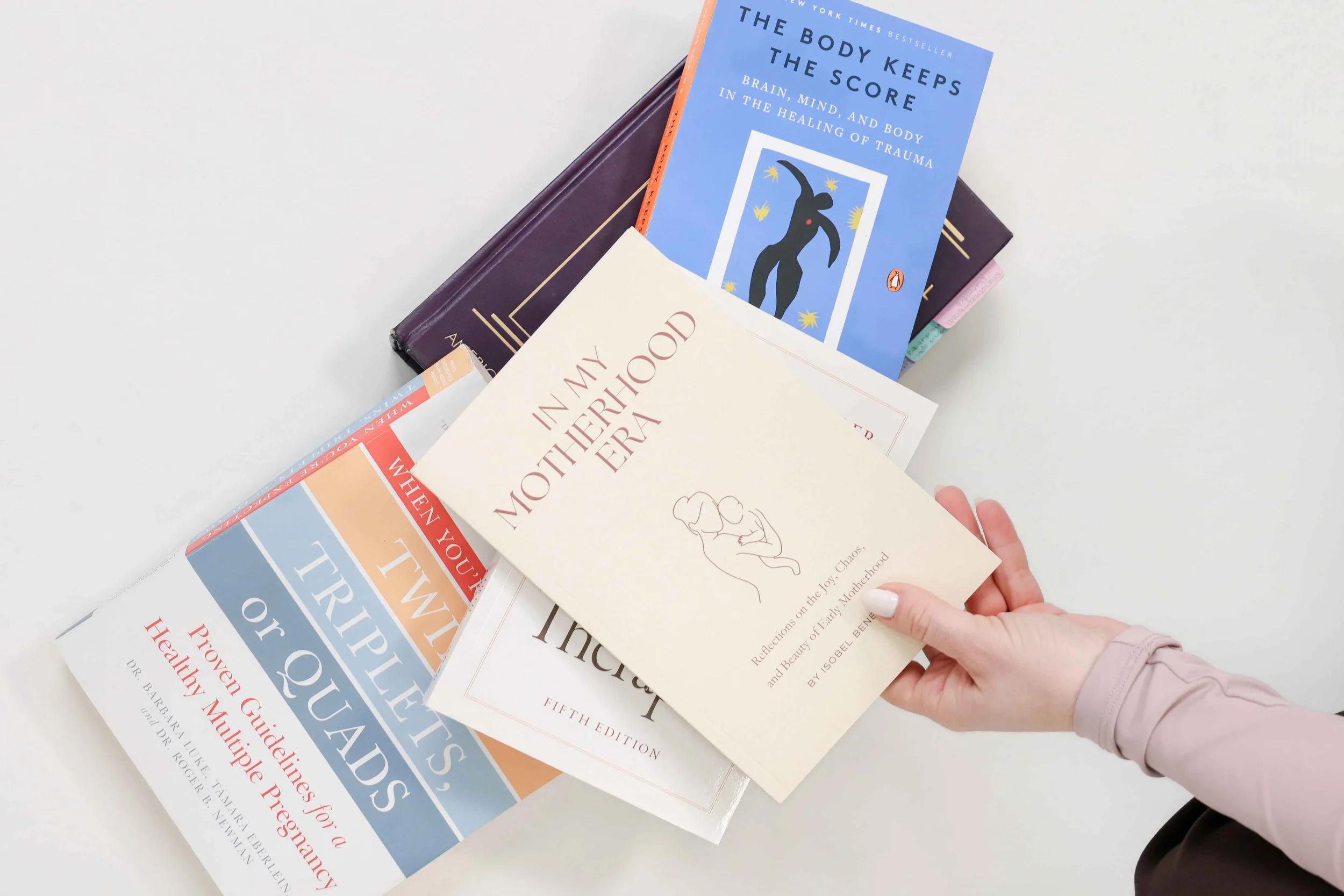 A person with light-colored nails, wearing a light pink jacket, is holding a paperback book titled "In My Motherhood Era" over a stack of several other books on a white surface. The stack includes a blue book titled "The Body Keeps the Score" and other books with colorful covers.