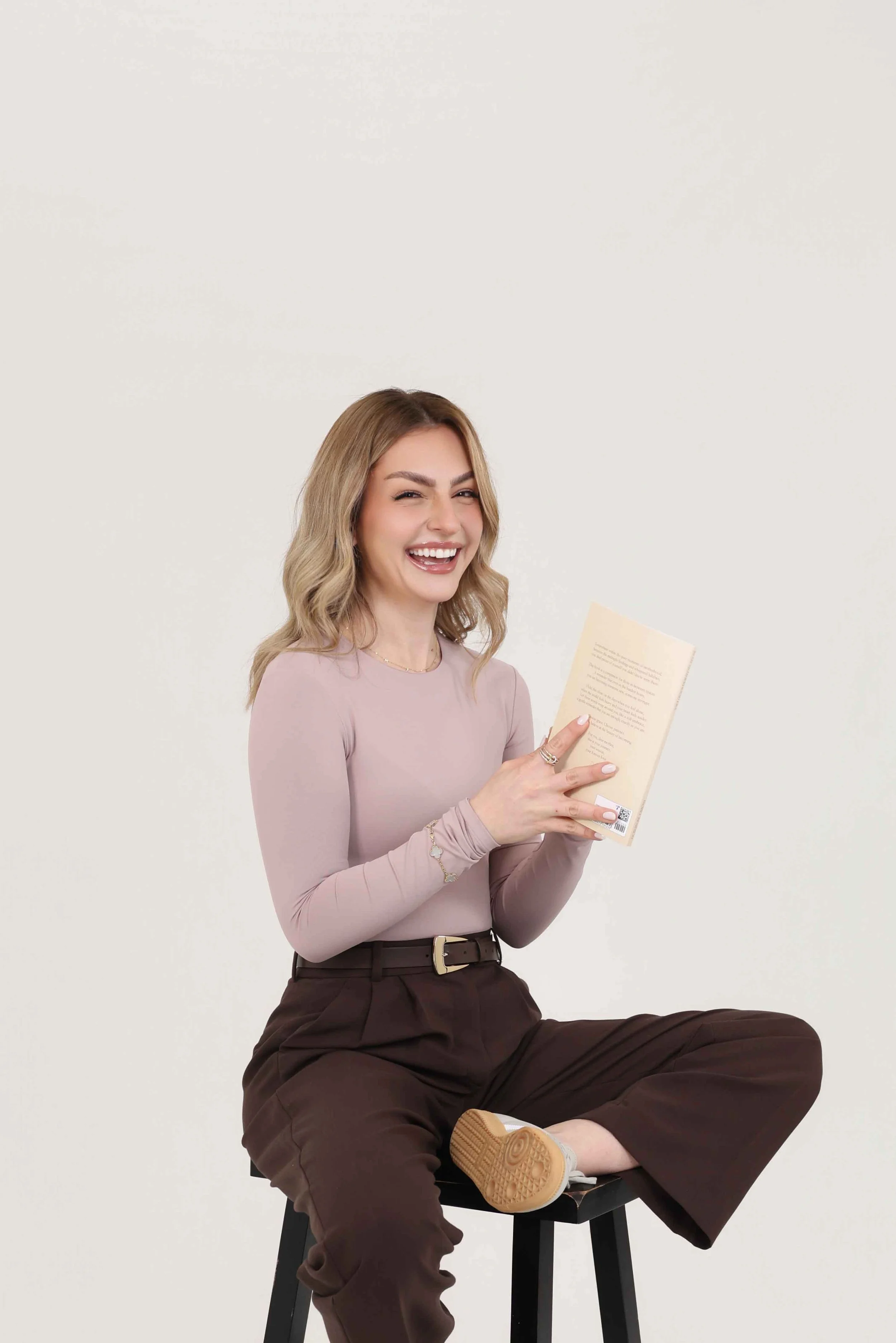 A young woman with blonde curly hair sitting cross-legged on a black stool, holding a paper and smiling at the camera against a plain white background.