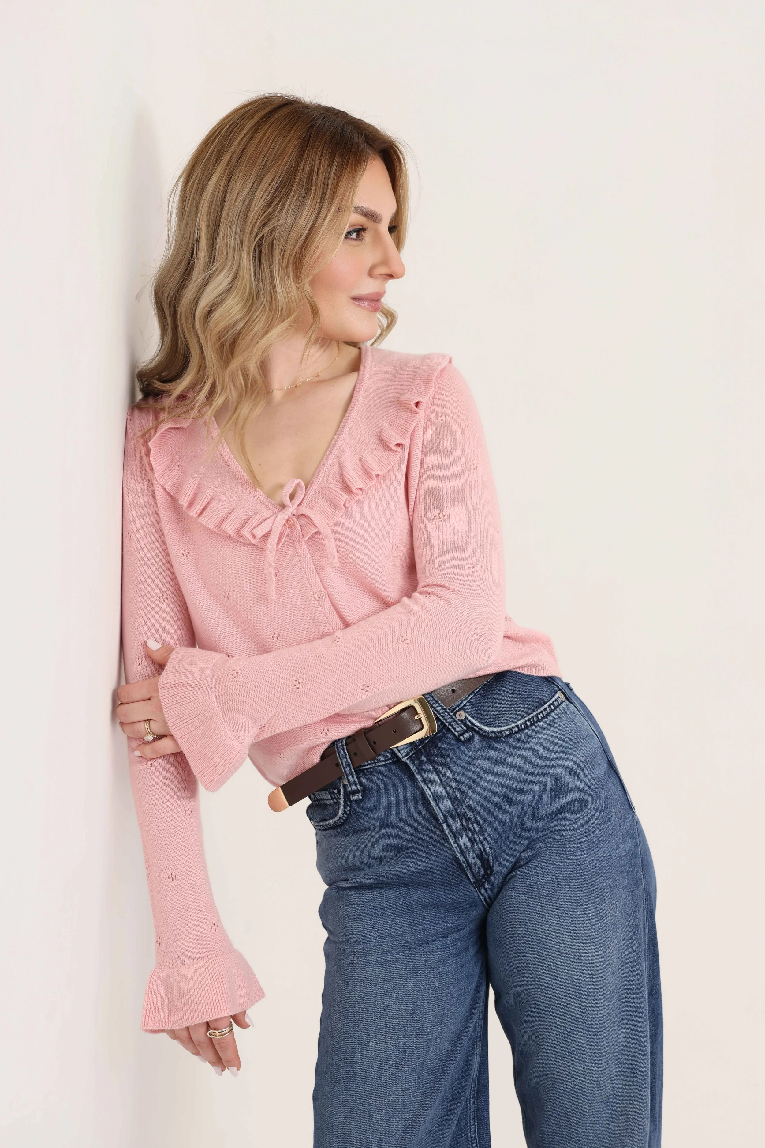 A woman wearing a pink ruffled sweater and blue jeans stands against a plain white background, looking to the side.