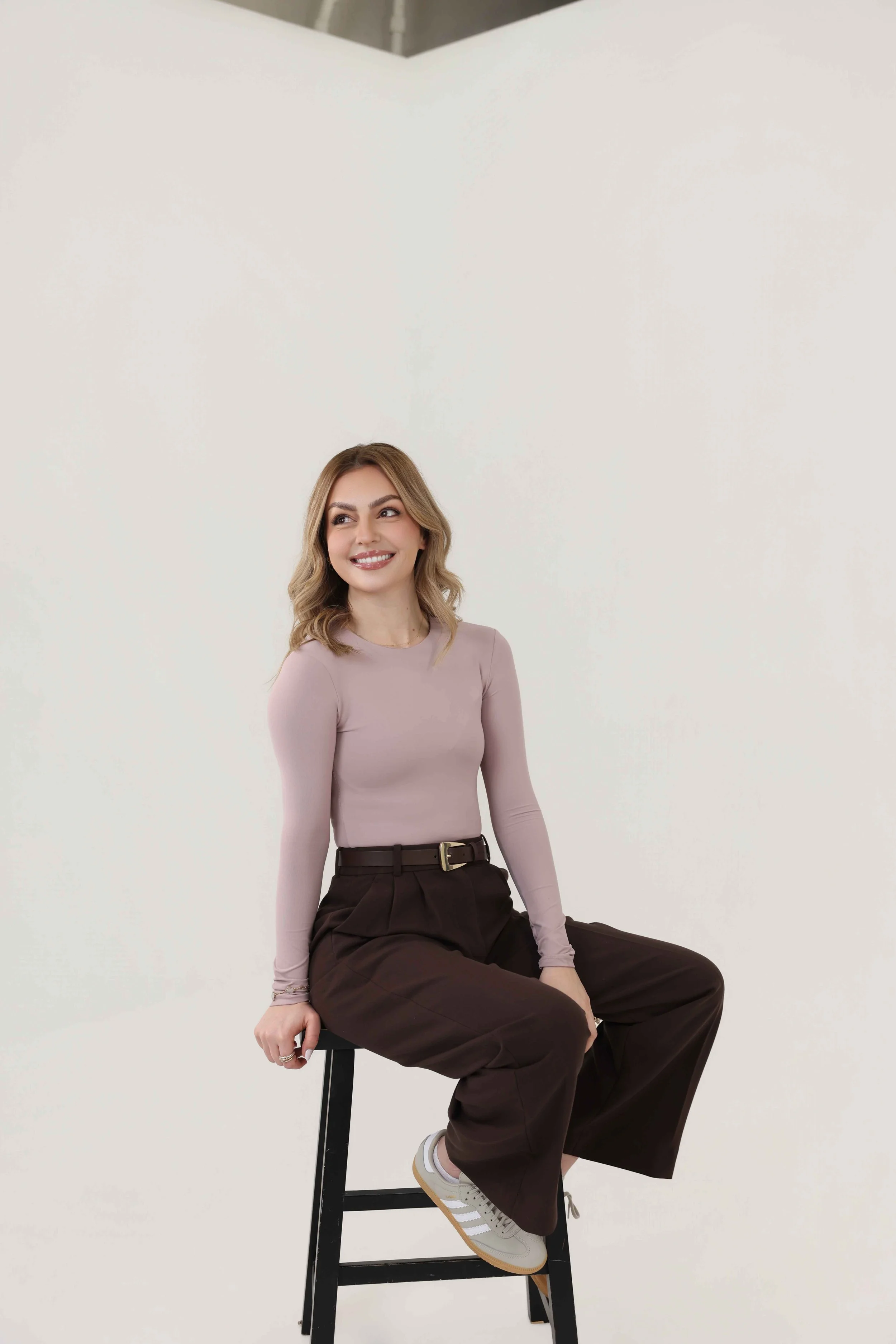 Young woman sitting on a black stool in a neutral-colored room, smiling and looking to her left. She wears a light pink long-sleeve top, brown high-waisted wide-leg trousers, white sneakers, and has shoulder-length blonde wavy hair.