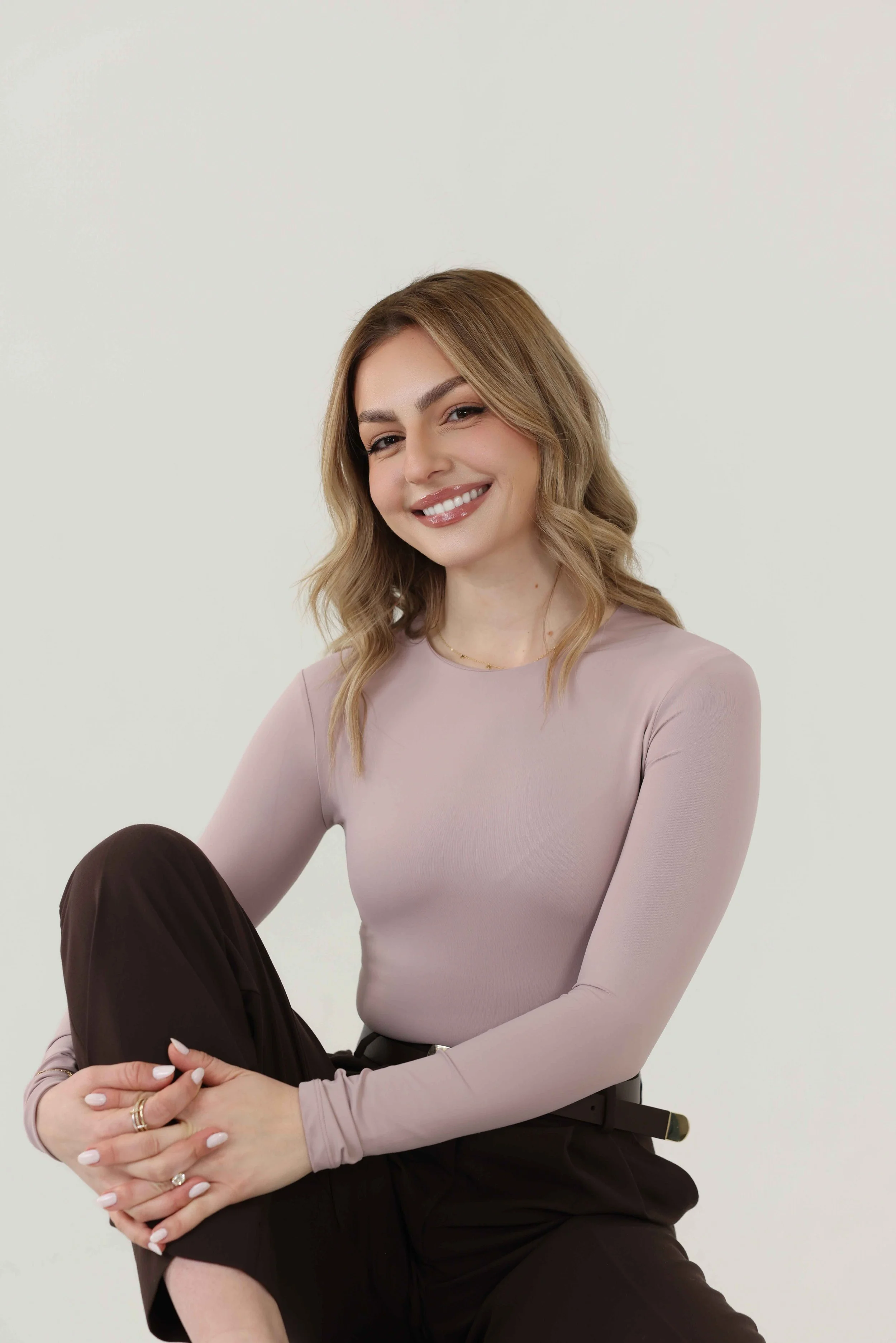 A woman with shoulder-length wavy blonde hair, wearing a light mauve long-sleeve top, dark pants, and jewelry, sitting against a plain white background with a warm smile.