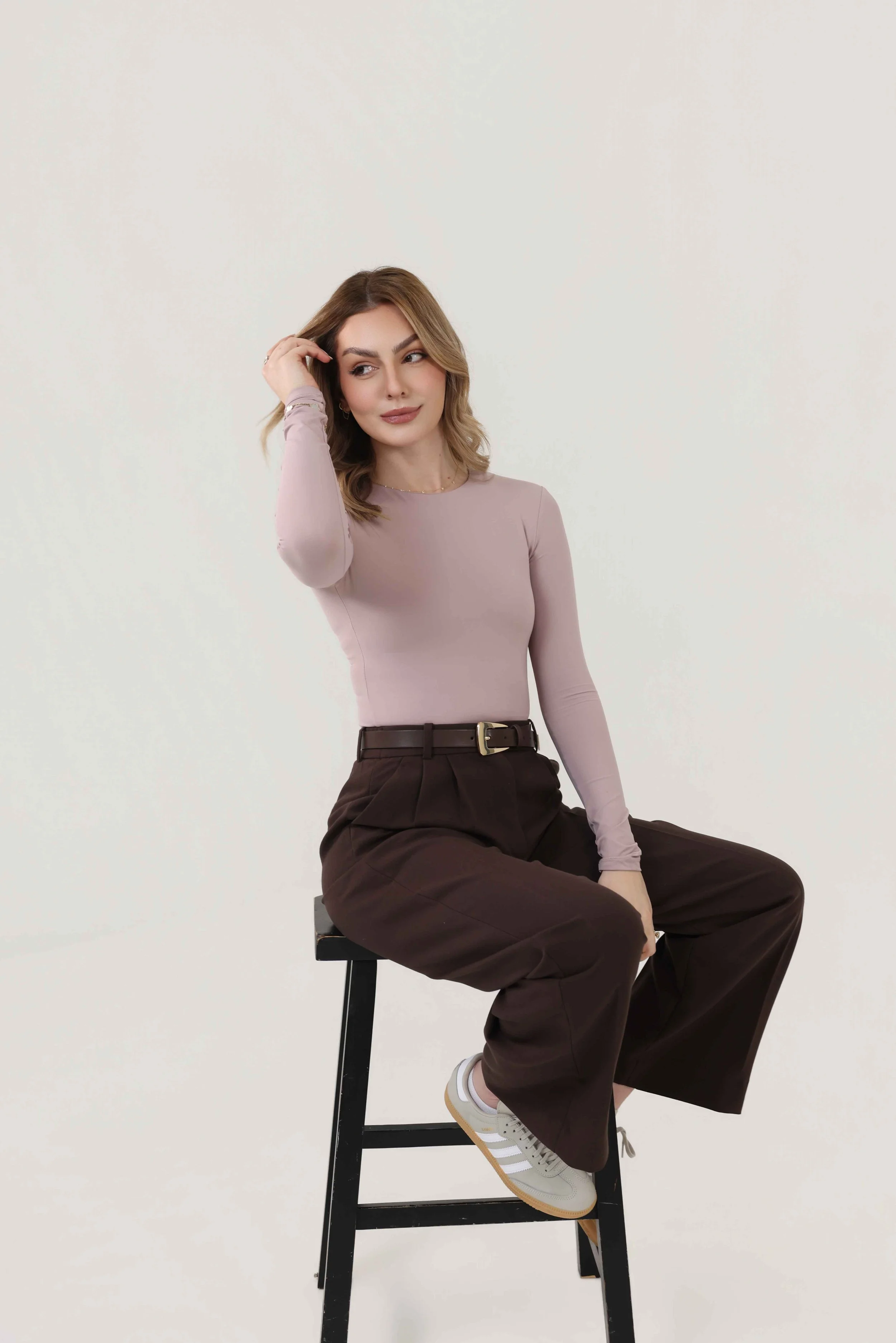 A woman with light brown hair sitting on a black stool against a plain white background. She is wearing a light pink long-sleeve shirt, brown high-waisted pants, white sneakers, and jewelry. She is touching her hair and looking to her left.