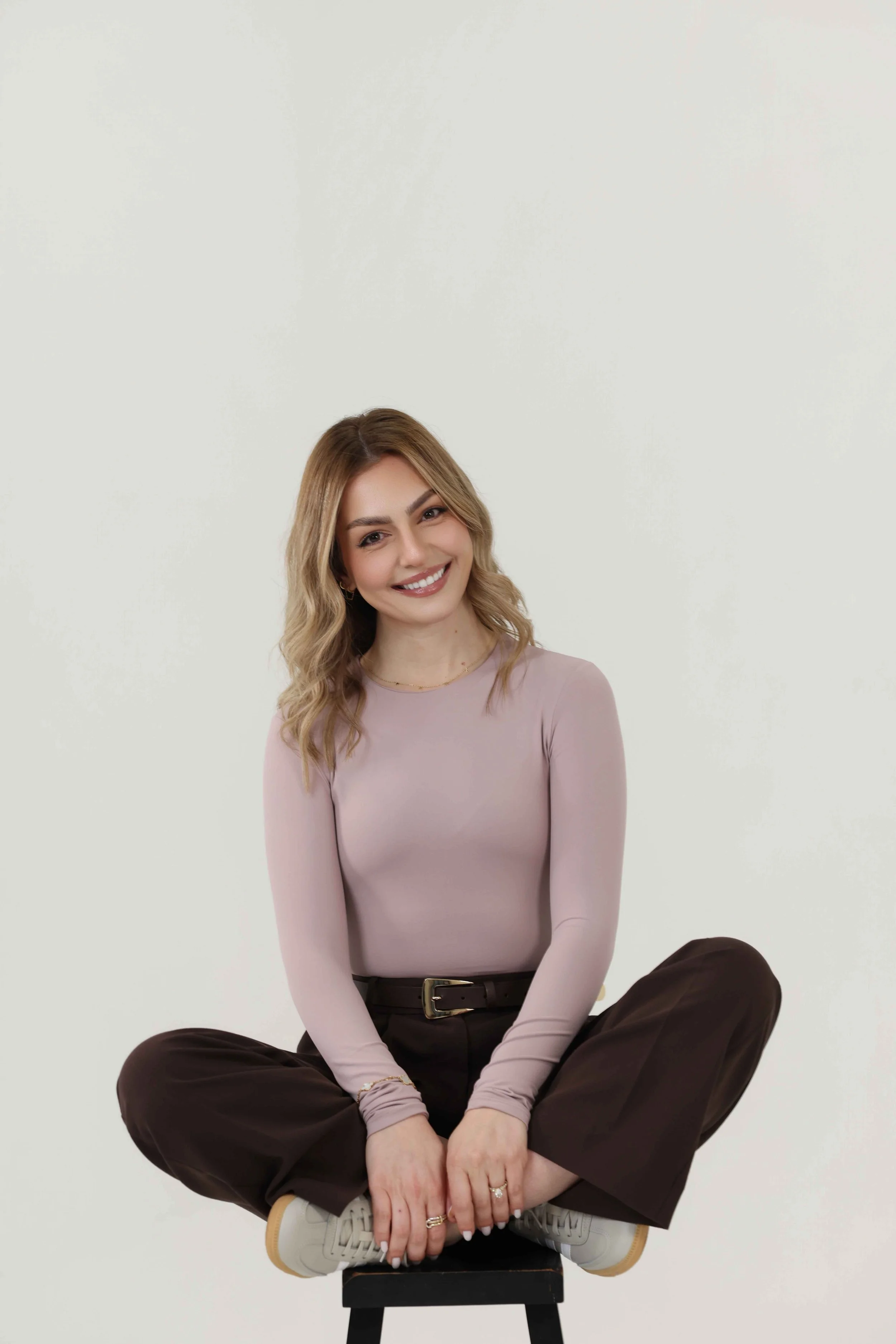 A smiling young woman with long wavy blonde hair, wearing a light pink long-sleeve top and dark brown pants, sitting cross-legged on a stool against a plain white background.