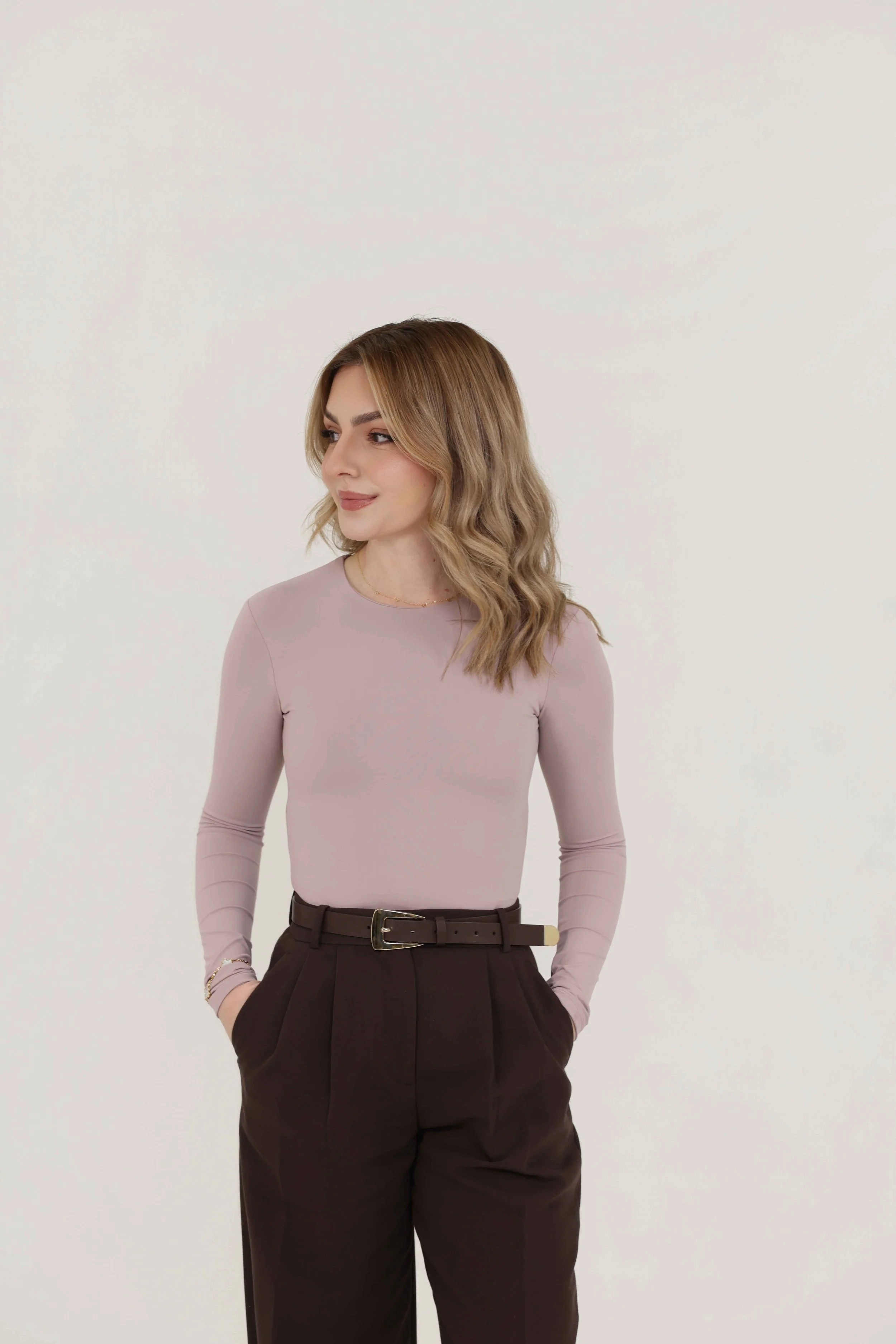 A woman with shoulder-length blonde hair styled in loose waves, wearing a light pink long-sleeve top and dark high-waisted trousers with a belt, standing against a plain off-white background.