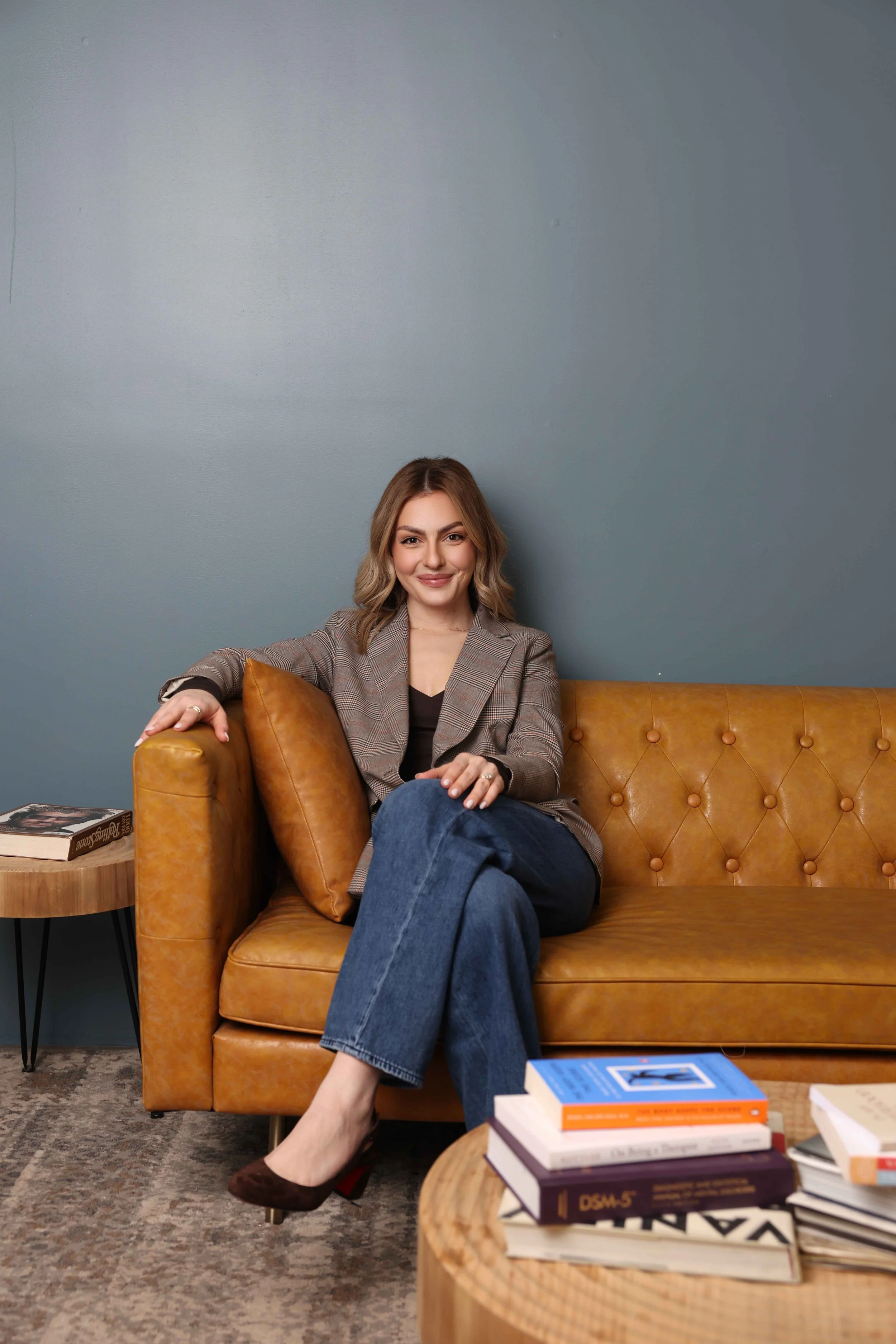 A woman sits on a tan leather couch with her legs crossed, smiling at the camera. She has shoulder-length blonde hair and is wearing a plaid blazer, a black top, and dark blue jeans. A pillow is next to her on the couch. In front of her, there is a wooden coffee table cluttered with books, some stacked and some scattered, with titles visible on the spines. To her left, there is a small round wooden side table with black metal legs, holding more books. The background features a gray wall, and the floor has a patterned rug.