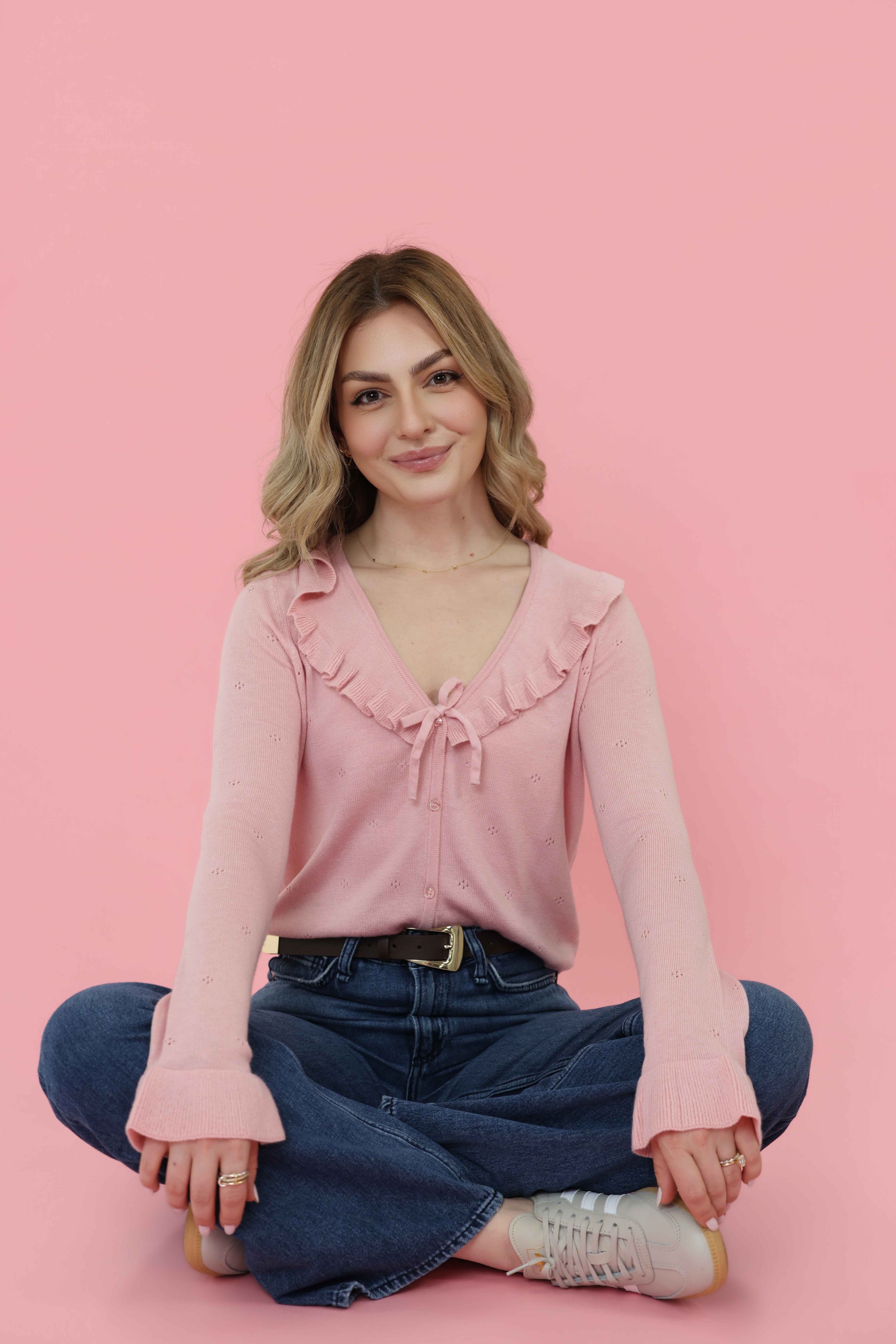 A woman sitting cross-legged against a pink background, wearing a light pink sweater with ruffles and a bow, dark blue jeans, white sneakers, and gold jewelry, smiling at the camera.