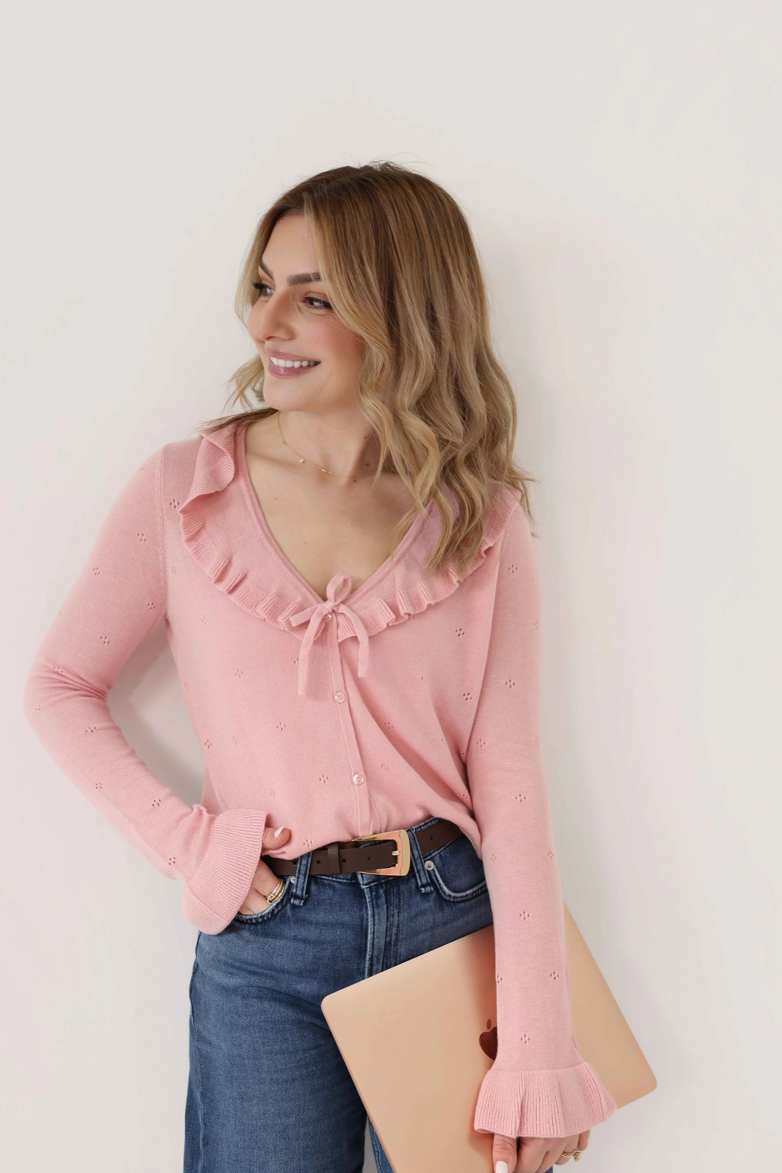 A woman wearing a pink sweater with ruffles and a bow, tied at the front, and dark jeans with a black belt, holding a rose gold MacBook, standing against a plain light-colored background.