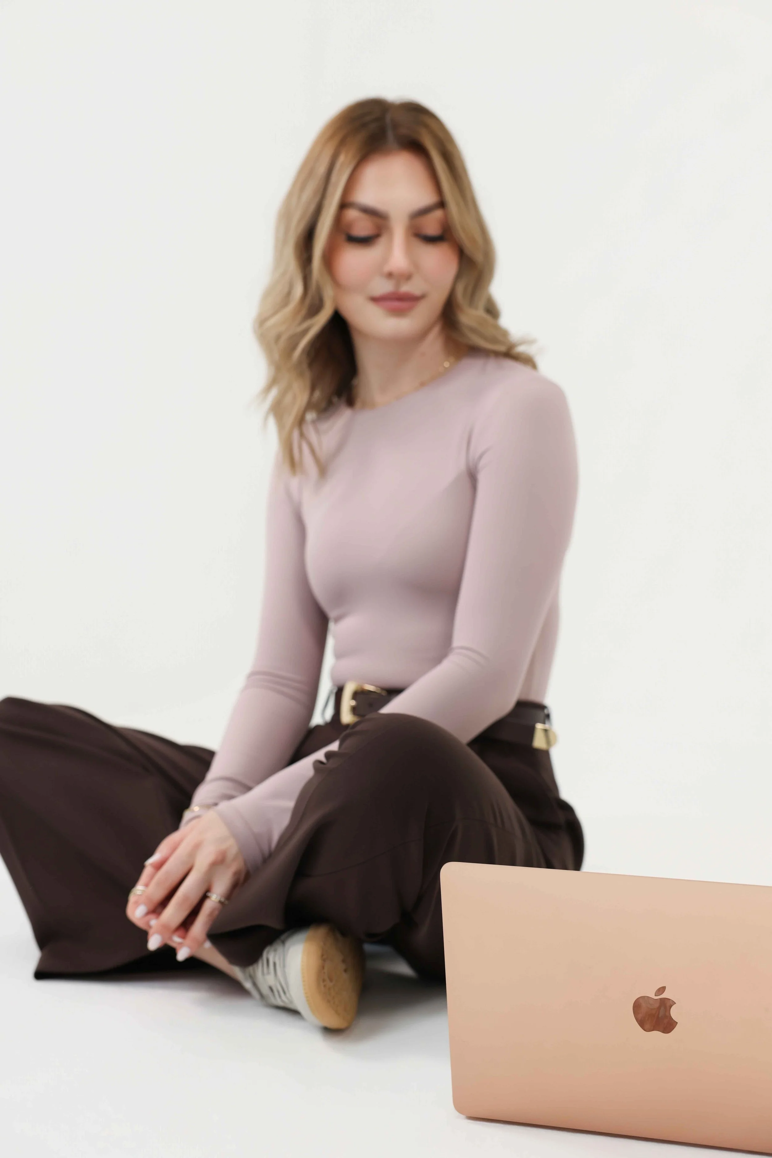 A woman with light brown hair and fair skin, dressed in beige and brown, is sitting with her eyes closed against a plain white backdrop. There is a gold Apple MacBook with a rose gold finish and Apple logo placed on the white surface in front of her.