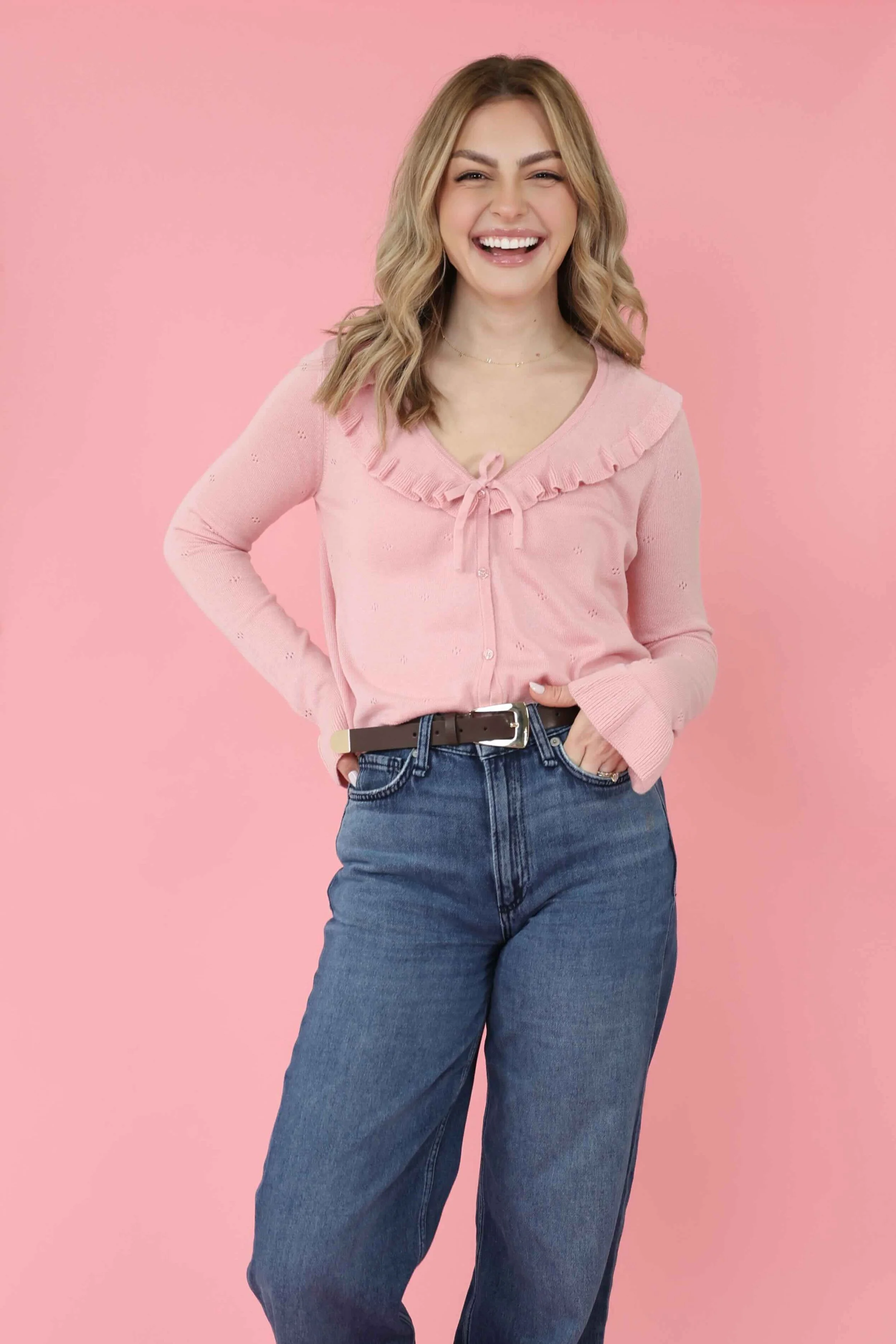 A woman with blonde hair styled in loose waves, wearing a pink long-sleeved sweater with ruffle details around the neckline, and blue jeans with a black belt. She is smiling and looking at the camera against a pink background.
