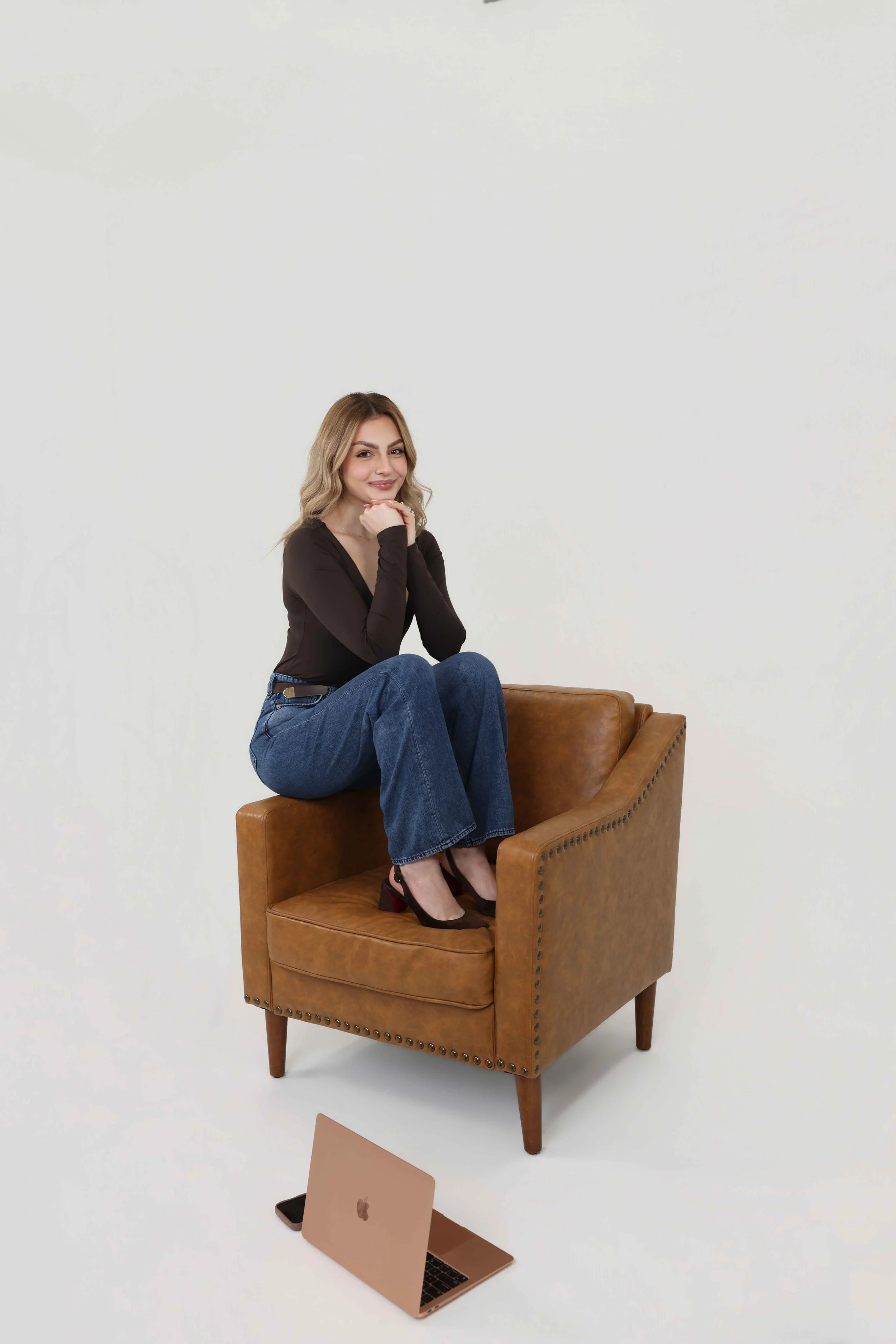 A woman sitting on an orange leather armchair, smiling and resting her chin on her hands, with a laptop, tablet, and smartphone on the floor in front of her, set against a plain white background.