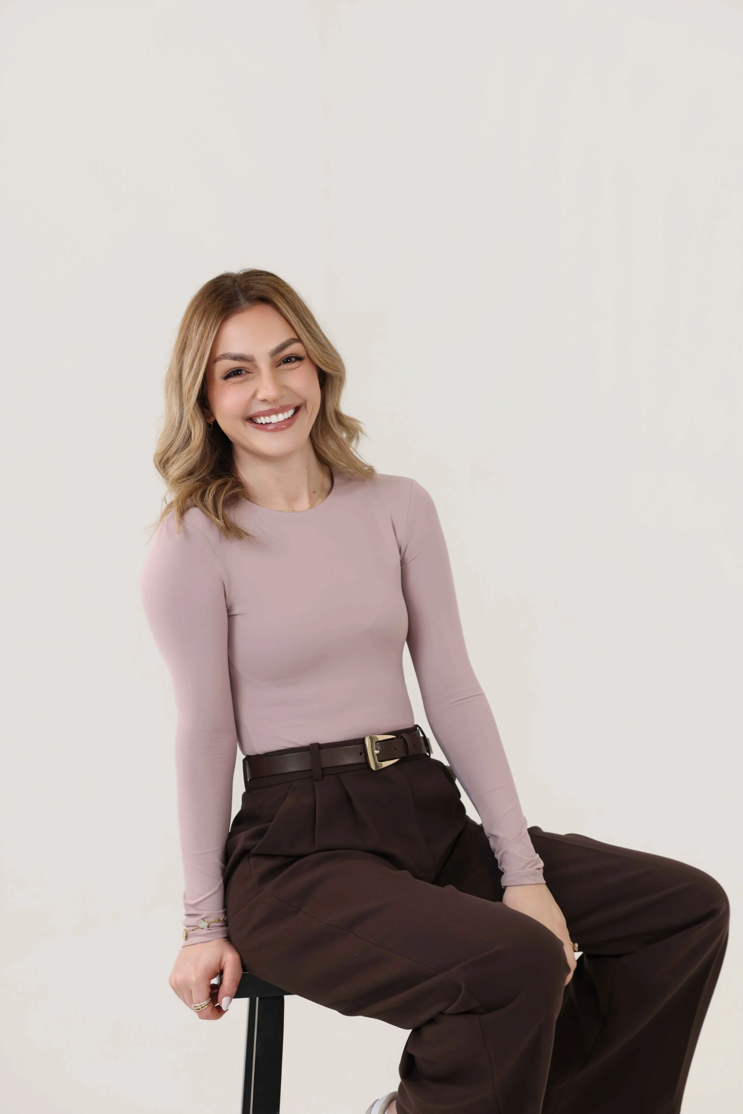Woman sitting on a black chair, smiling, against a plain white background, wearing a light pink long-sleeve top and dark brown pants with a belt.