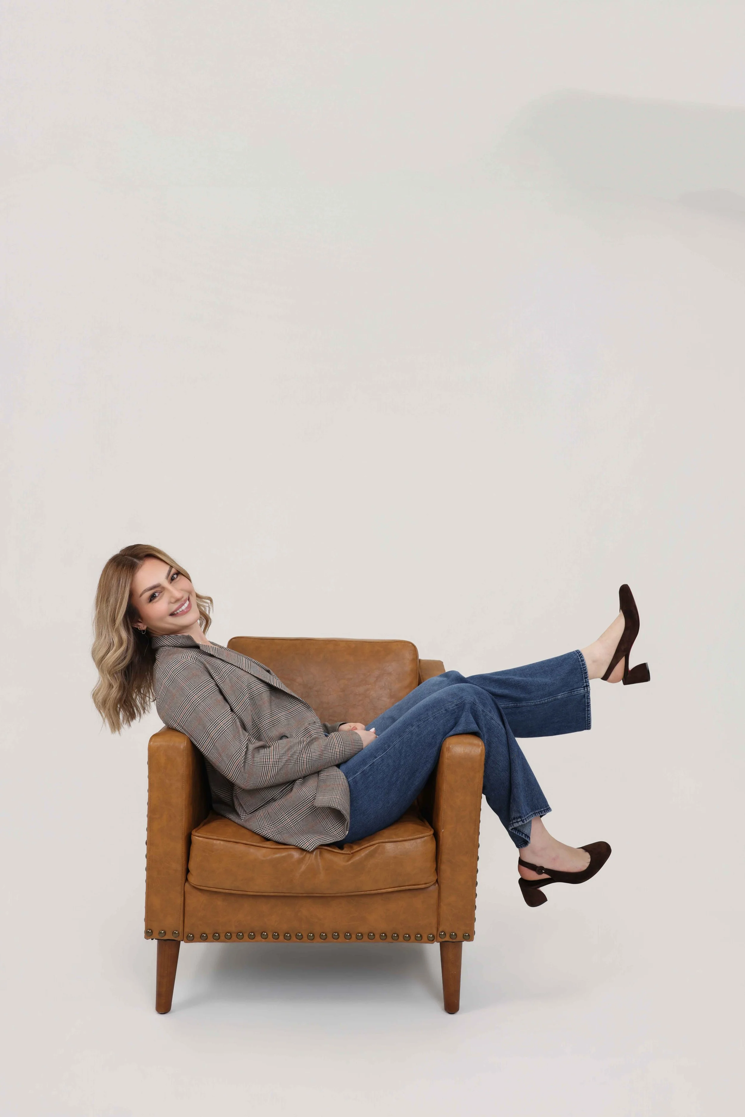 Woman with blonde hair smiling, wearing a plaid blazer and blue jeans, reclining with her legs resting on the arm of a brown leather armchair, and wearing brown heels, set against a plain light background.
