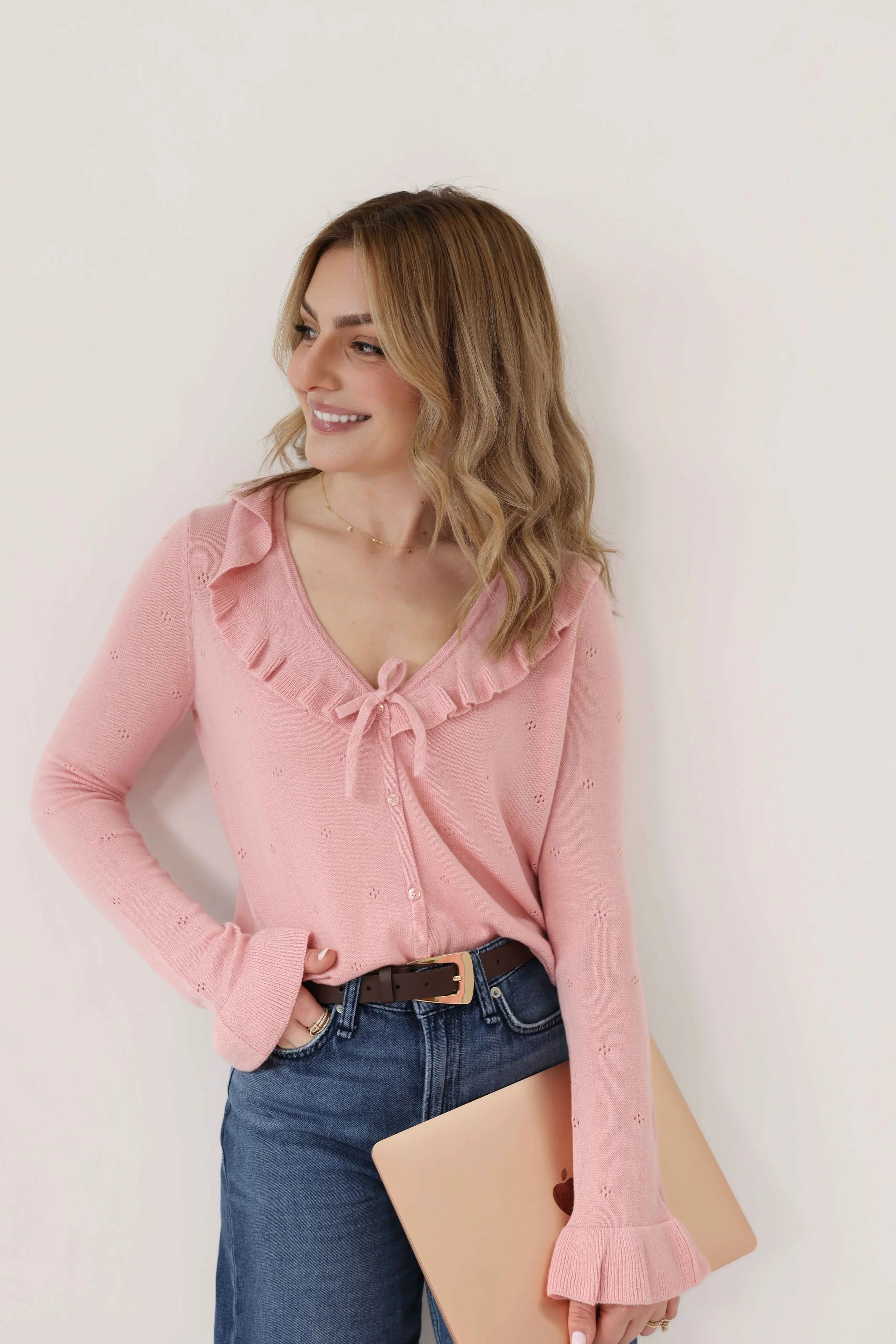 Woman with blonde hair and a pink cardigan, smiling, standing against a white background with a laptop in her hand.