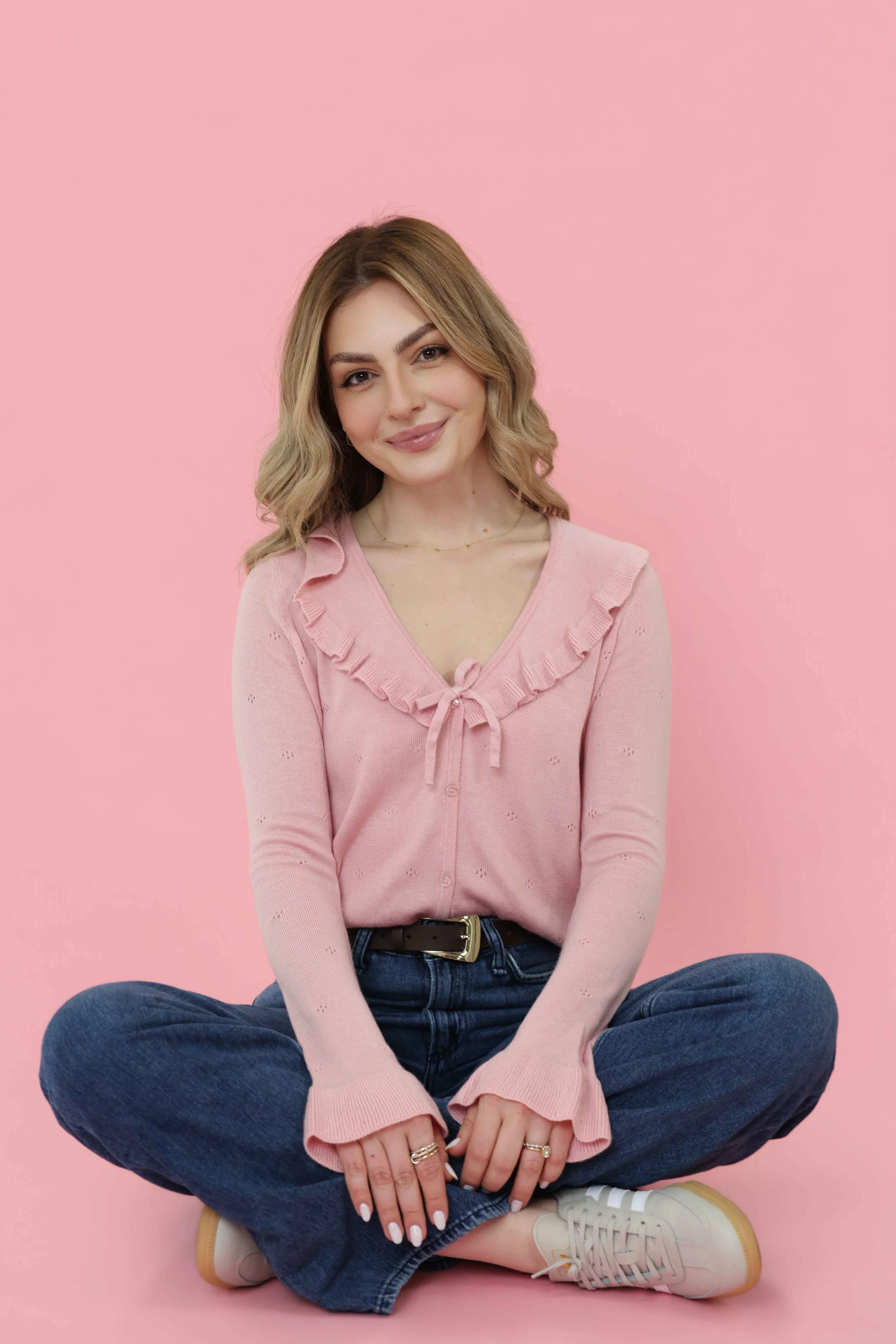 A young woman with shoulder-length wavy blonde hair sitting cross-legged against a pink background. She is wearing a light pink sweater with ruffled details and a bow at the neck, dark blue jeans, and white sneakers. She is smiling softly and looking directly at the camera.