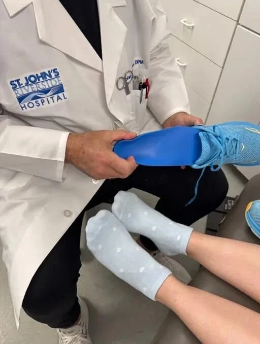 A doctor in a white coat at St. John’s Riverside Hospital holds a bright blue orthopedic shoe, while a patient wears socks and sits on an examination table.