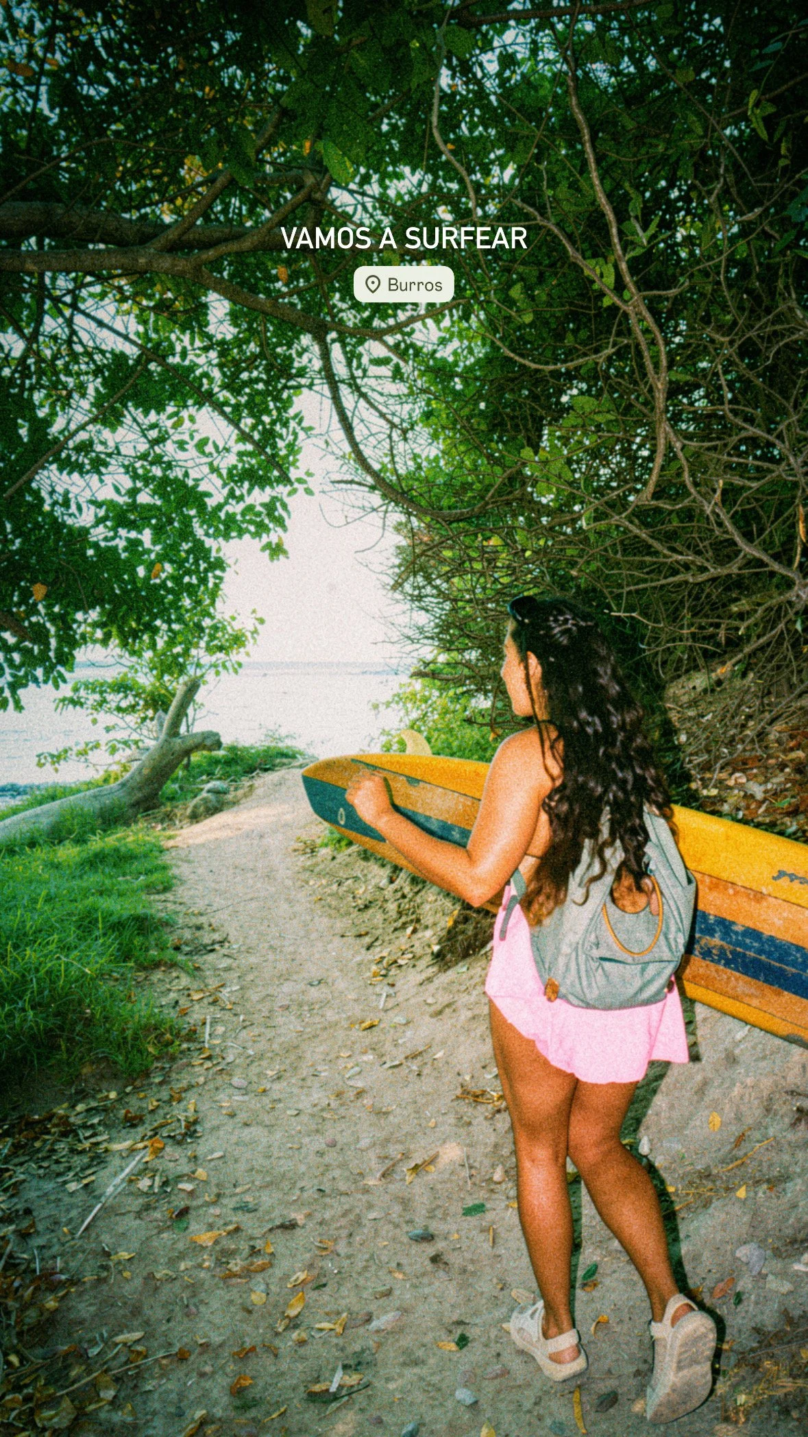 A young woman with long curly hair holding a surfboard, walking along a wooded trail near a body of water, with trees and green foliage surrounding her.