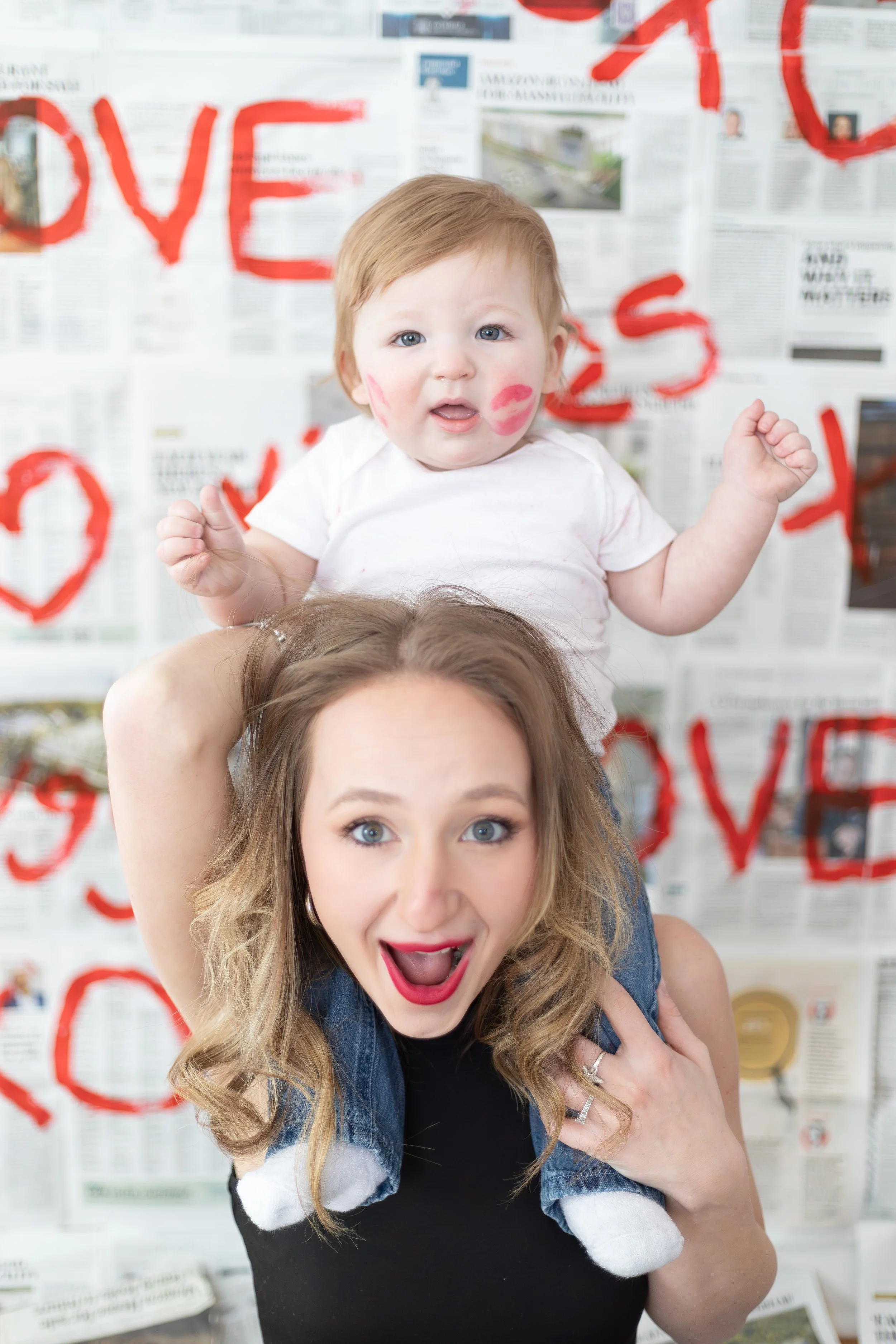 A woman with long wavy blonde hair making a surprised face and smiling as she holds a young child with red hair and blue eyes, who has pink lipstick marks on their cheeks and is sitting on her shoulders against a background of newspaper pages with re