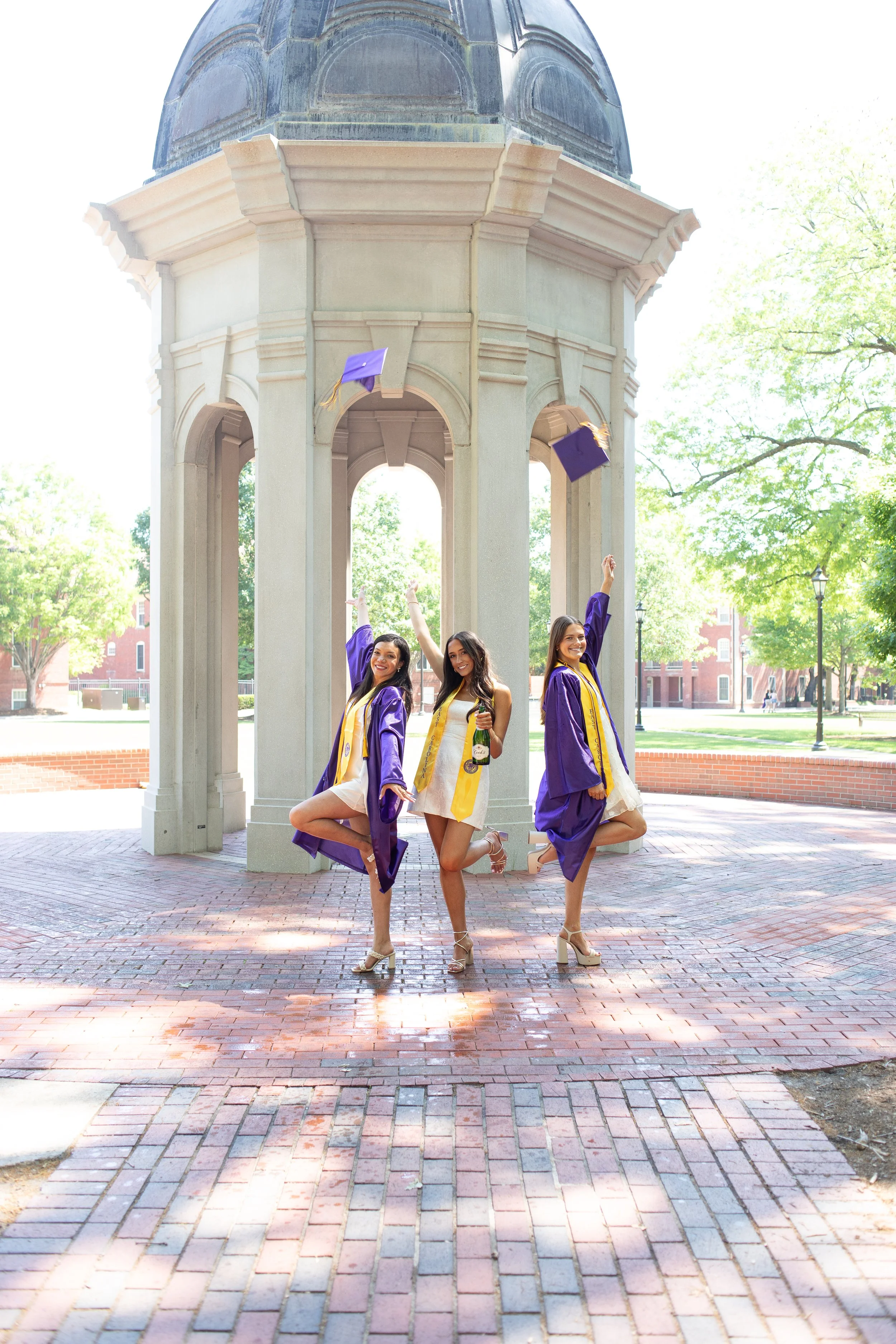 Three women in graduation gowns and caps celebrating outdoors under a pavilion, with trees and buildings in the background.