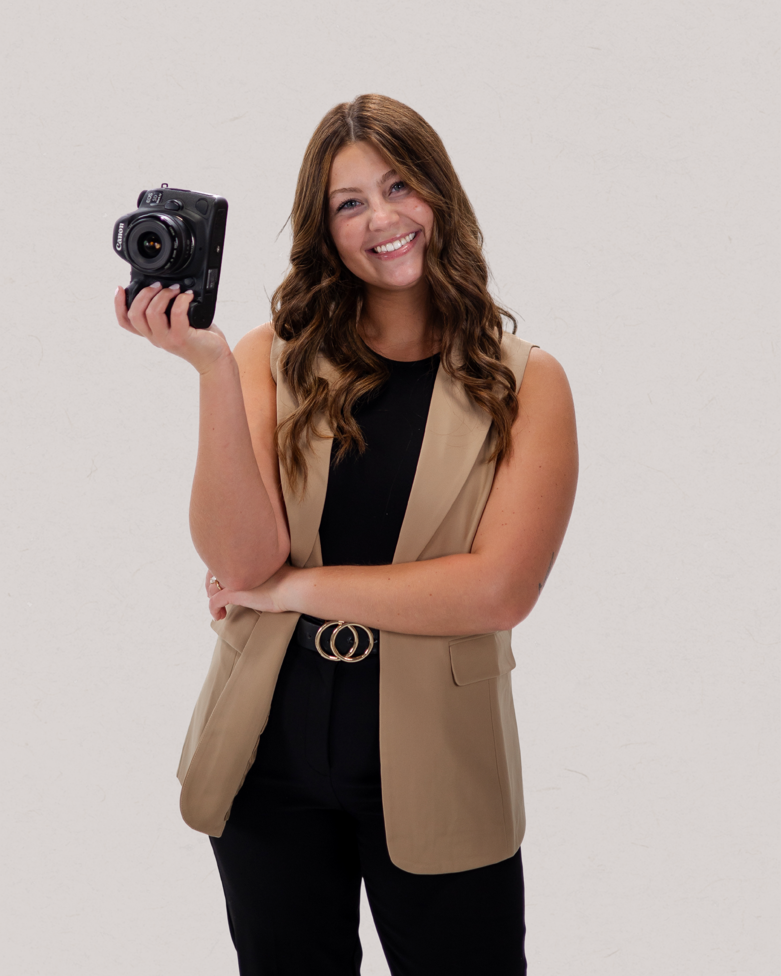 A woman with long wavy brown hair smiling and holding a professional camera in front of a plain white background.