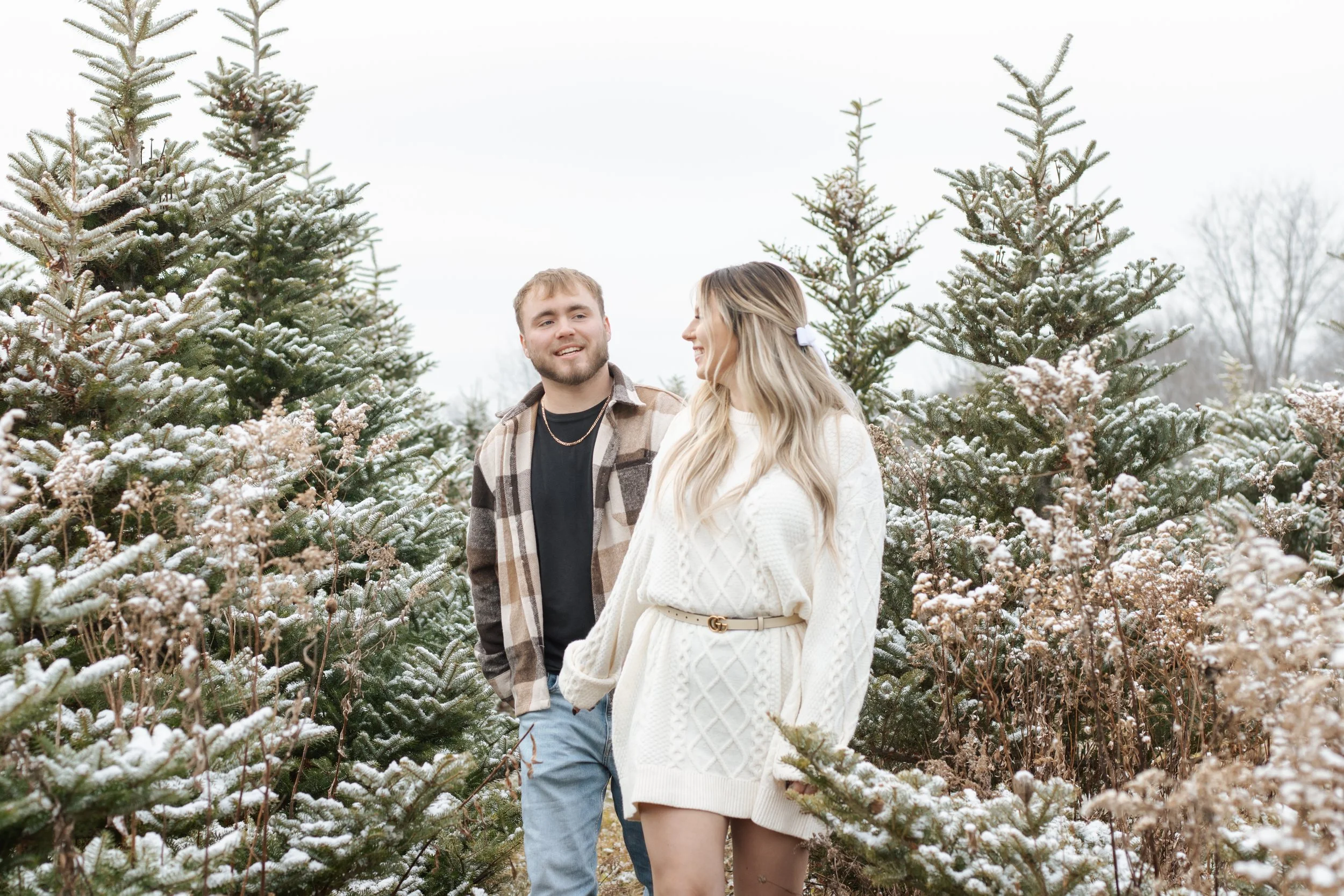 A young man and woman are walking through a snowy forest of pine trees, smiling at each other.