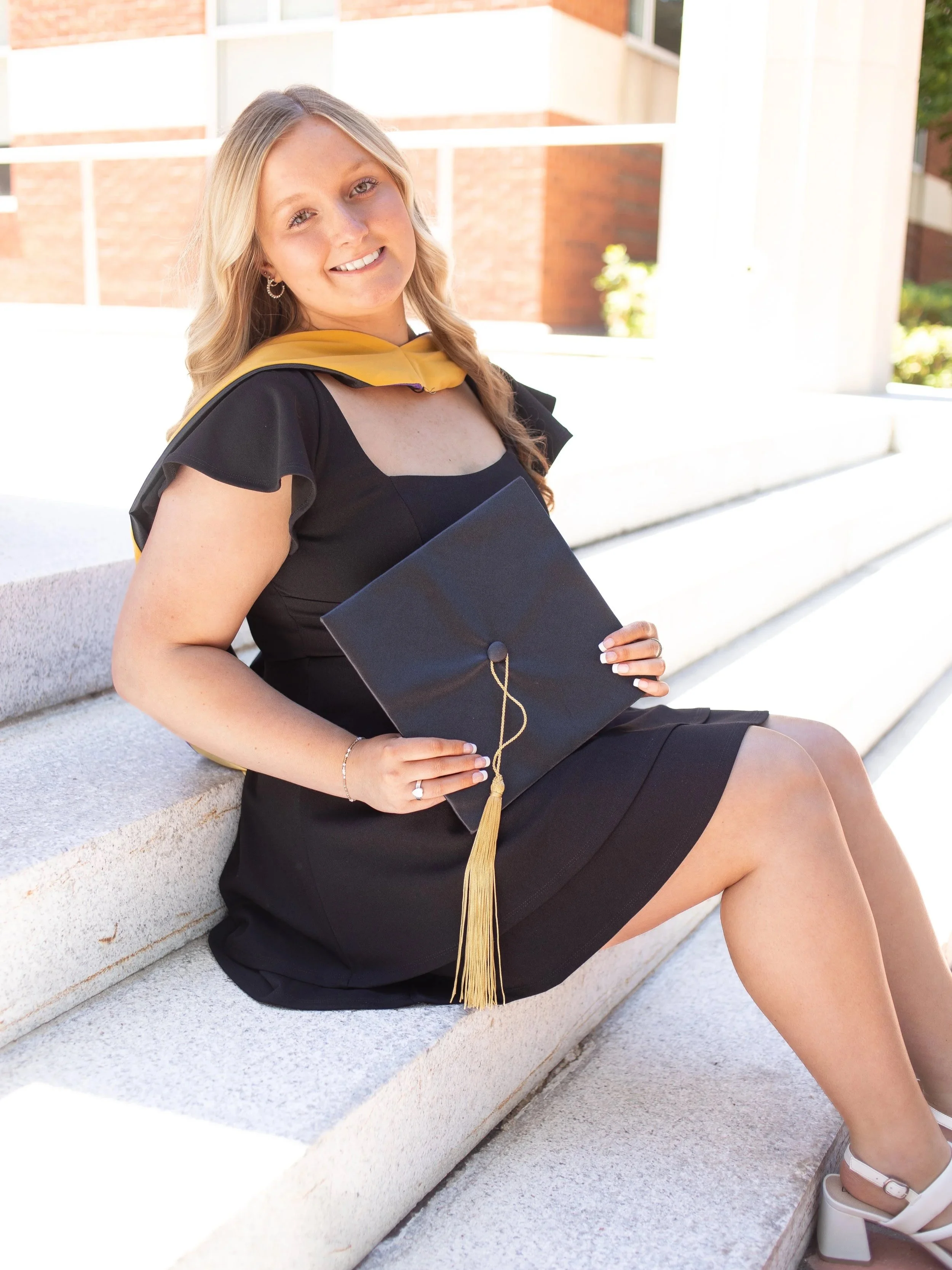 A young woman sitting on outdoor concrete steps dressed in a black dress with ruffled sleeves, holding a graduation cap with a gold tassel, smiling at the camera.