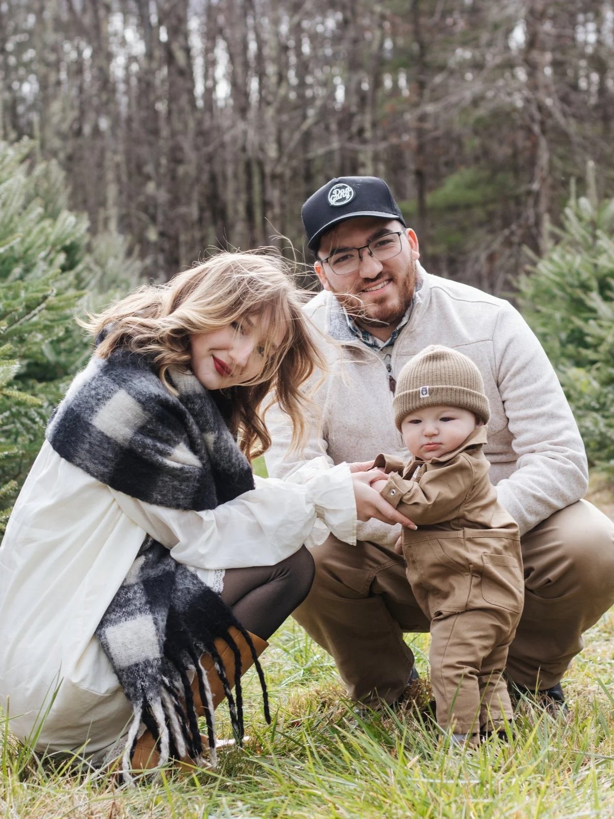 A family of three outdoors in a forested area with trees, a woman with wavy hair and a scarf, a man with glasses and a cap, and a toddler in a tan jacket and hat, holding hands.