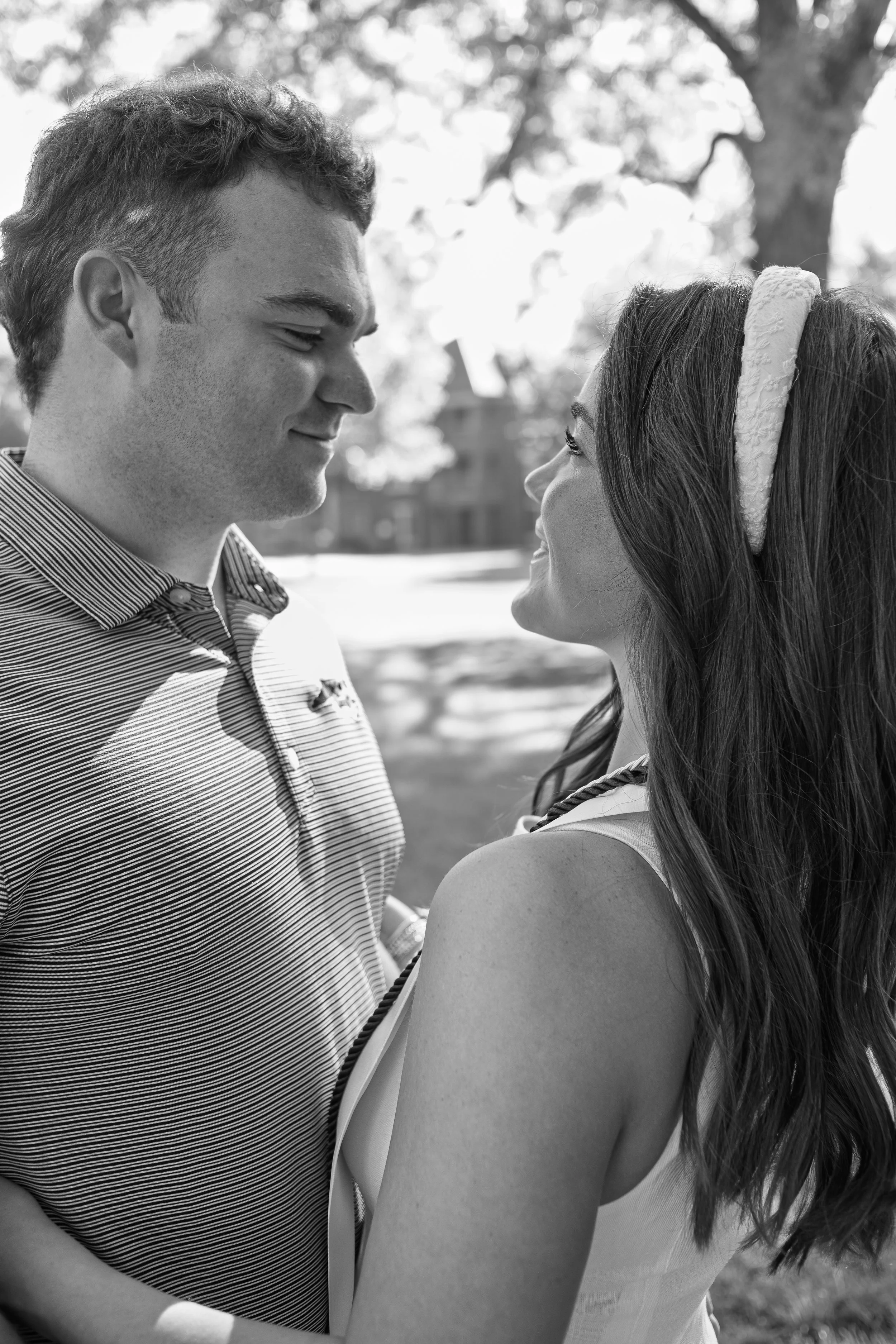 A black-and-white photo of a couple standing close, looking into each other's eyes outdoors with trees and sunlight in the background.