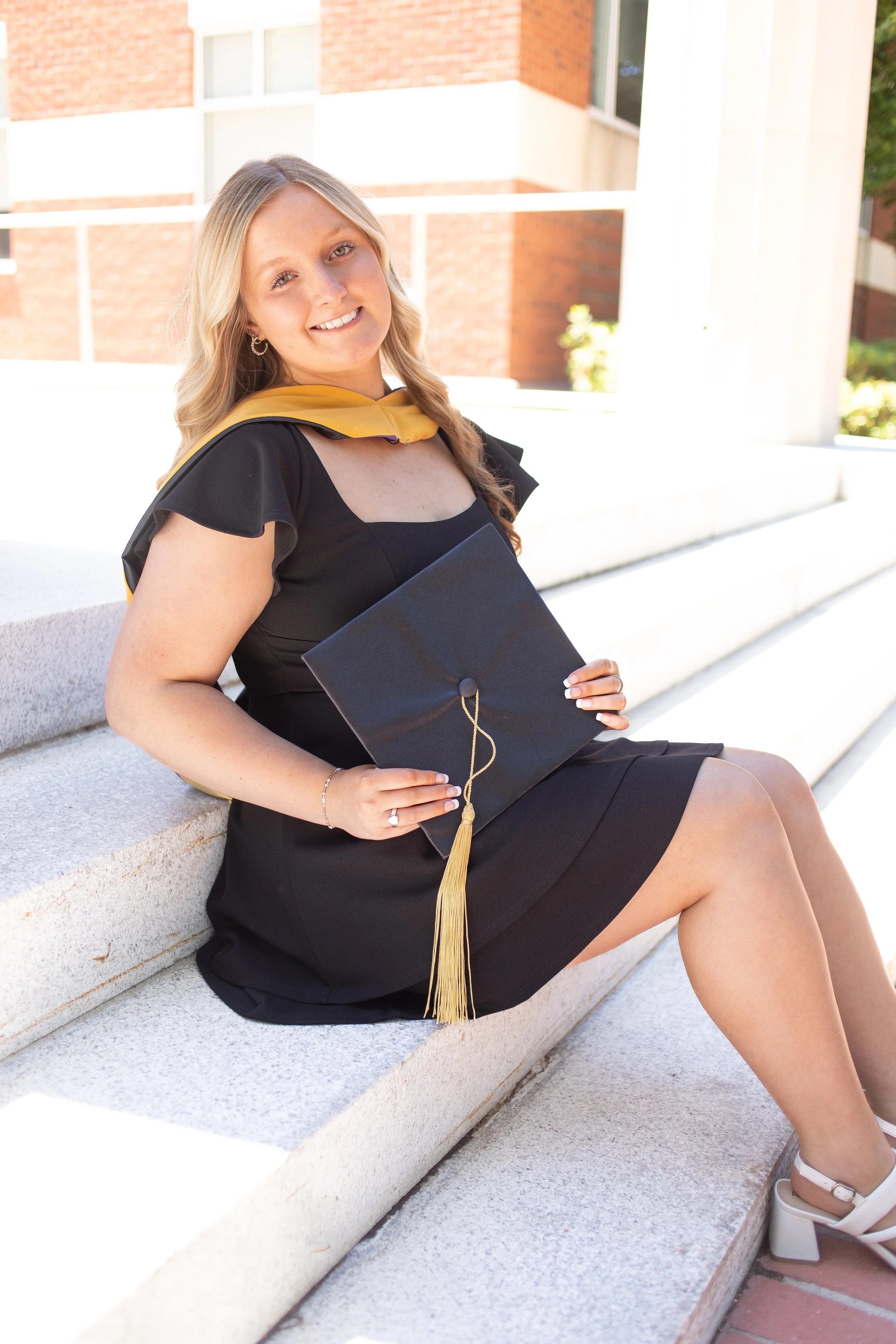 A young woman in a black dress sitting on outdoor steps, wearing a yellow stole and holding a black graduation cap with a gold tassel, smiling at the camera.