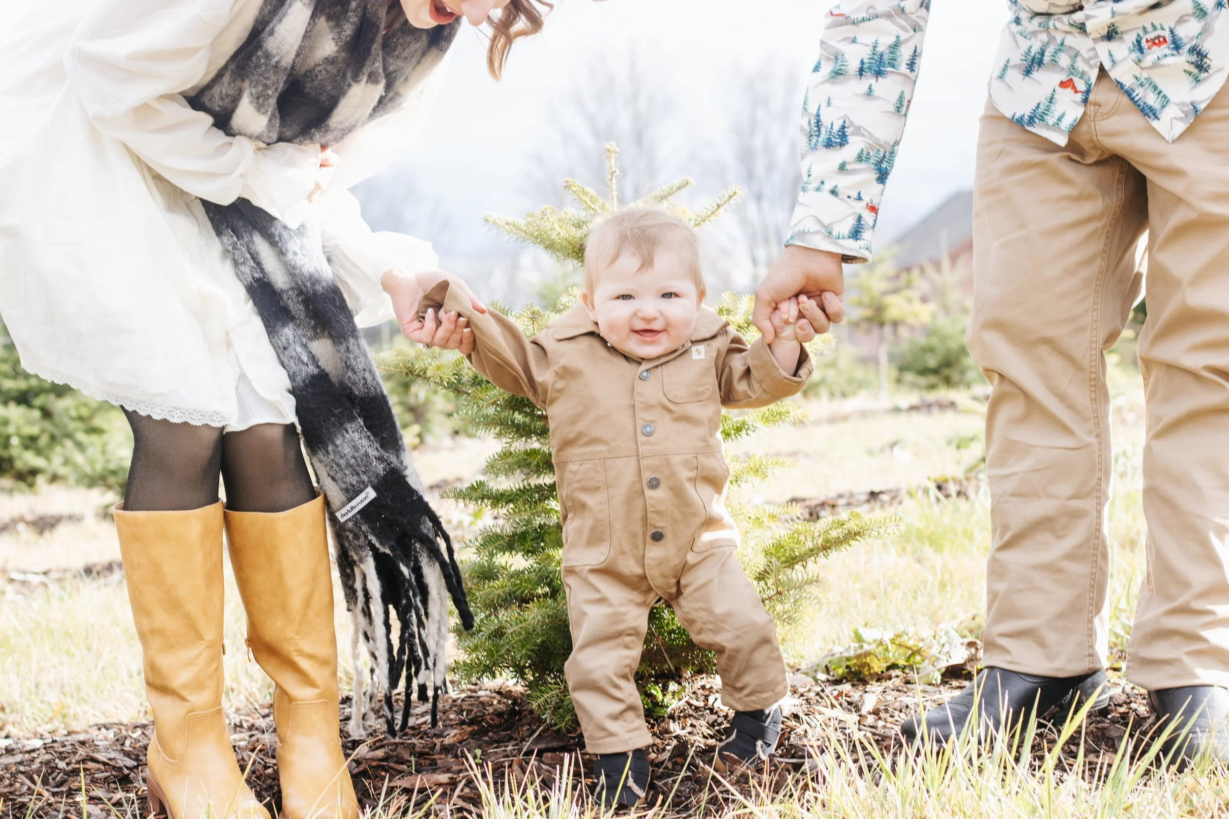 A young child holding hands with two adults, standing in a garden with a small Christmas tree. The child wears a brown jumpsuit and black boots. The adult on the left wears yellow rain boots and a white dress with a black and white scarf. The adult o