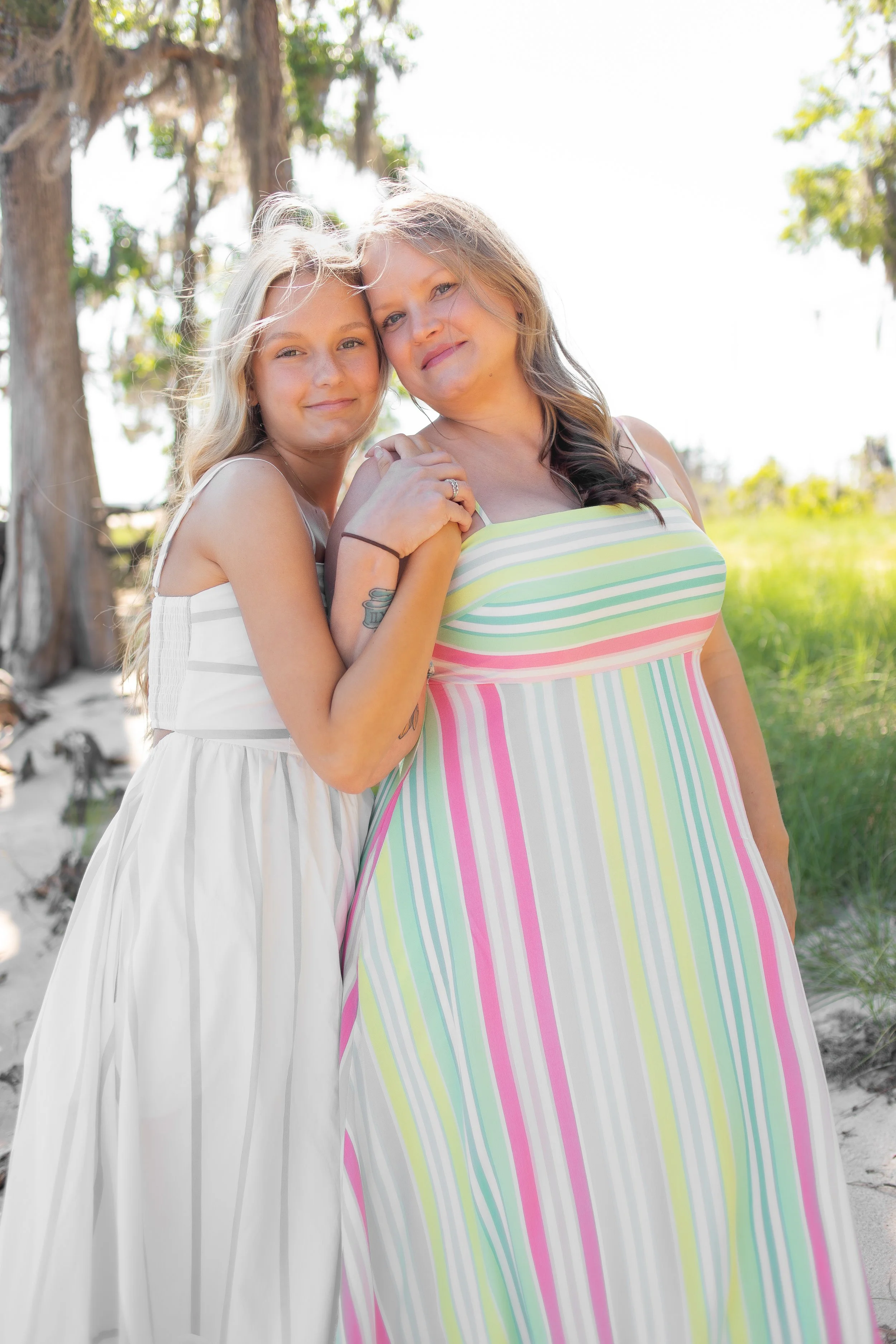 A woman and a girl hugging outdoors, with trees in the background and bright sunlight.