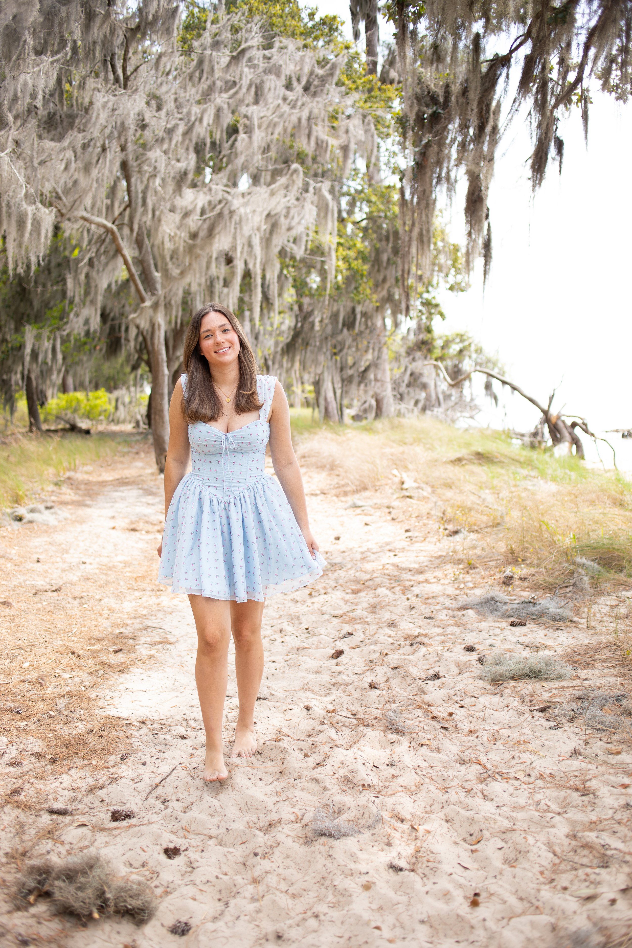 A young woman with long brown hair wearing a white sundress with small floral patterns, smiling, walking barefoot down a sandy trail surrounded by moss-draped trees.