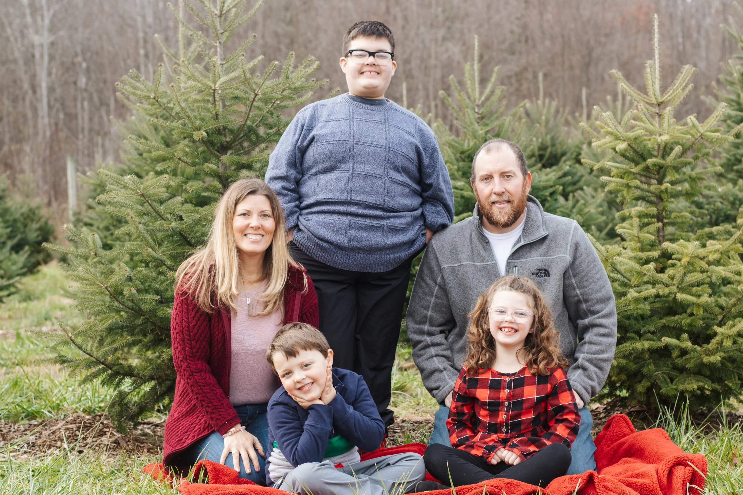 A family of six posing together outdoors during the fall, surrounded by evergreen trees, with some of them sitting on a red blanket on the grass. They are smiling and appear happy.