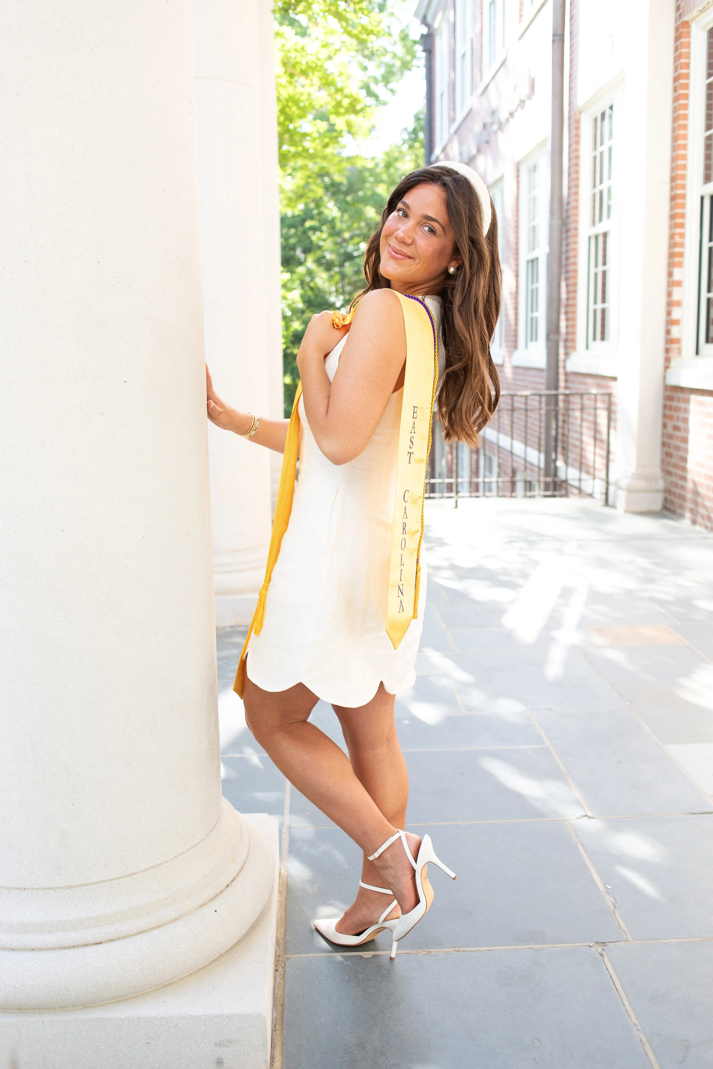 A young woman in a white dress and white high heels standing outdoors next to a building with brick and white columns, smiling and looking over her shoulder.
