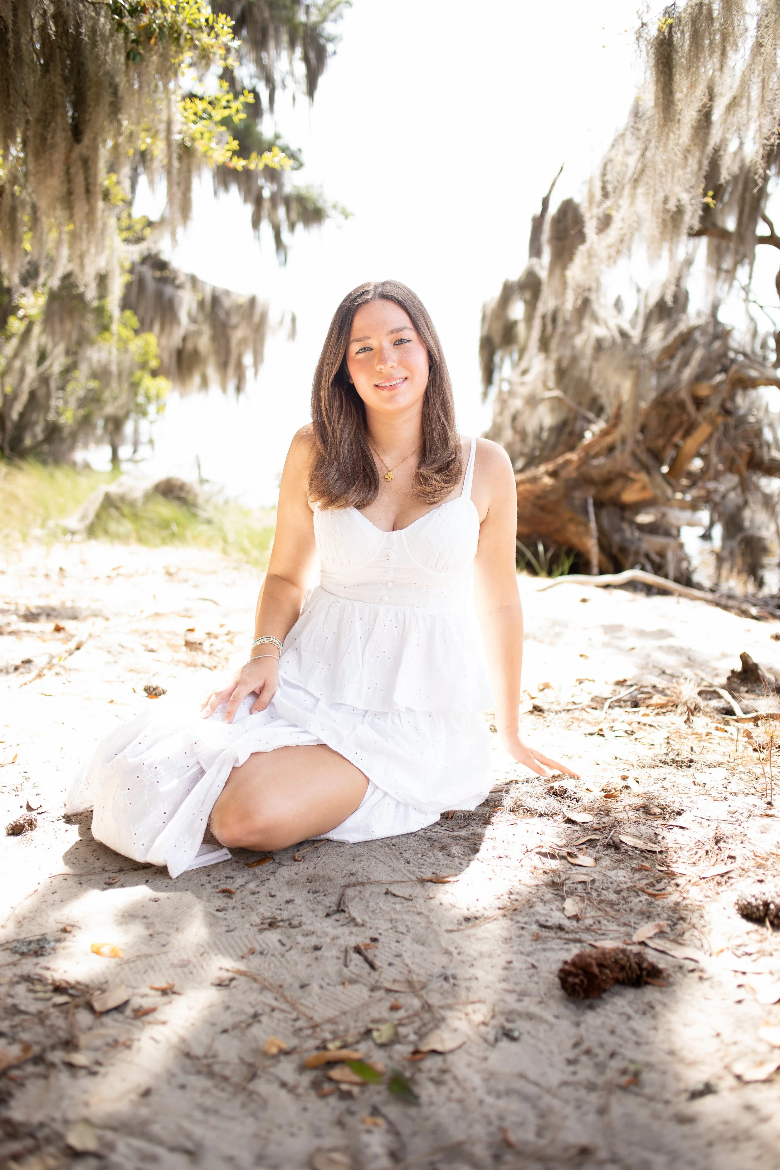 A woman in a white dress sitting on the sandy beach with trees draped in Spanish moss in the background.