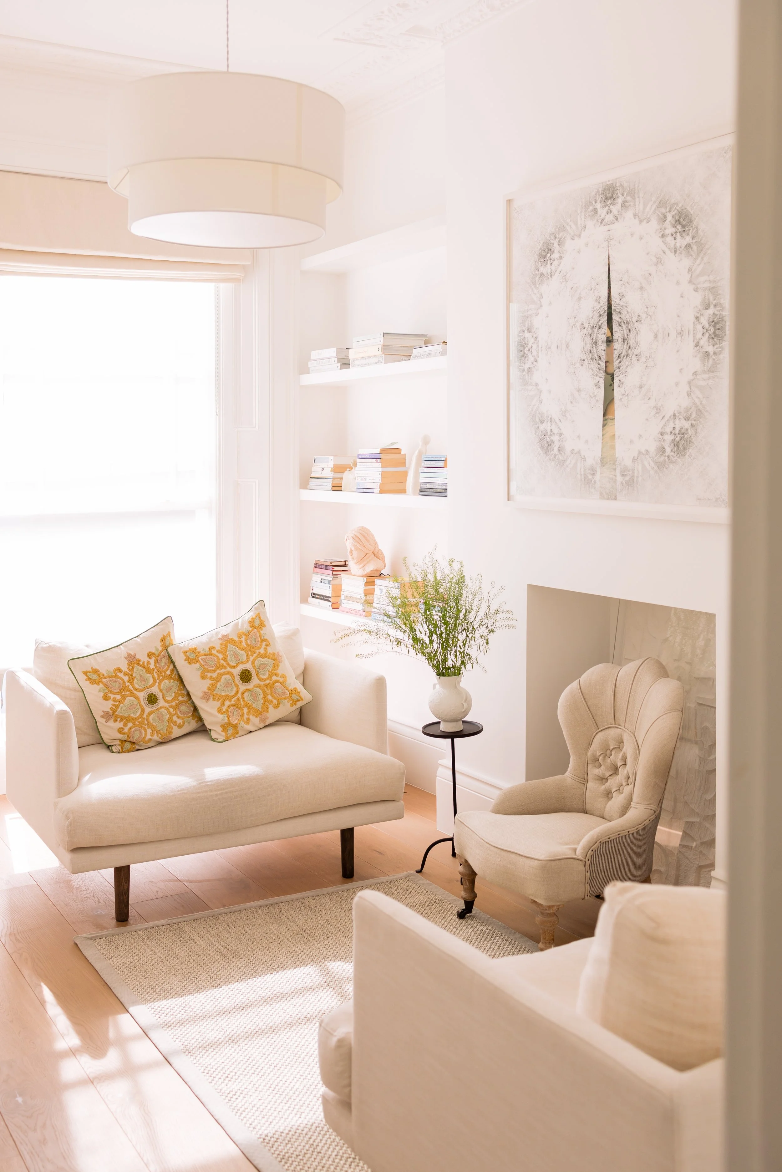 Bright and airy St John's wood consulting room with white sofas and armchairs, decorative pillows, a large abstract painting, a bookshelf with books, a potted plant, and a light-colored area rug on wooden flooring.