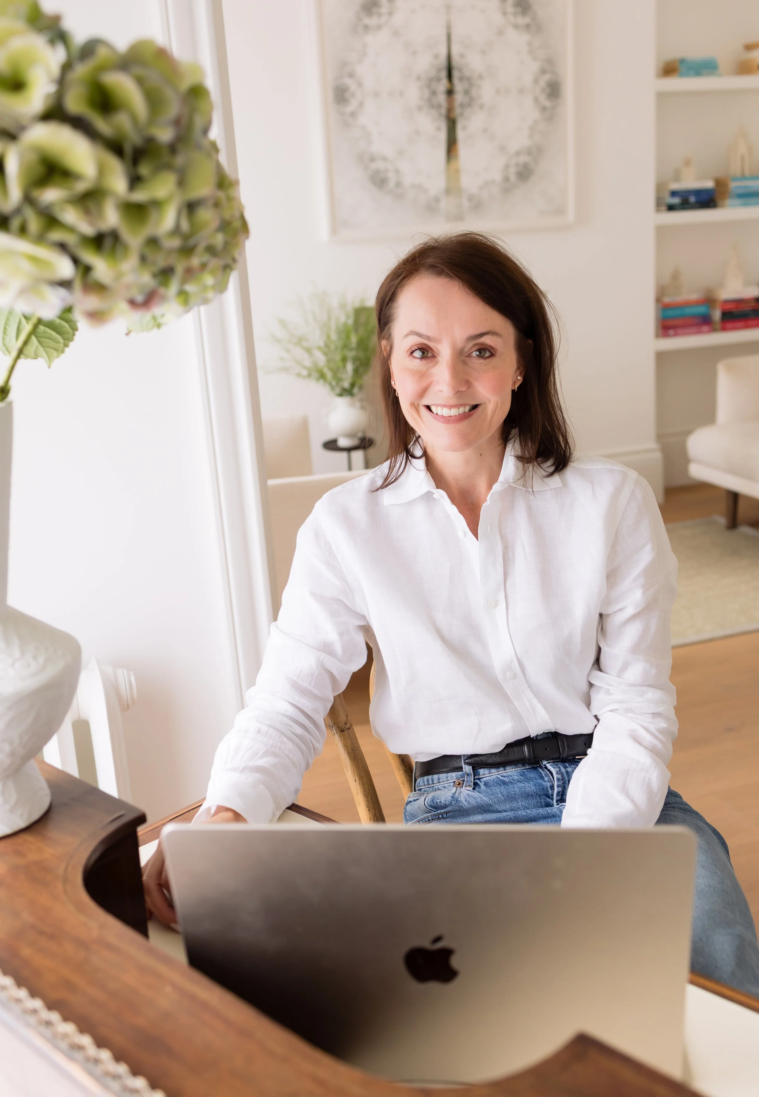 Jeanne Vorster Naidoo, Clinical Psychologist, smiling warmly as she sits at an antique writing desk with a laptop in her St John's Wood consulting room, wearing a white linen shirt and jeans, with green hydrangeas in the foreground and bookshelves an