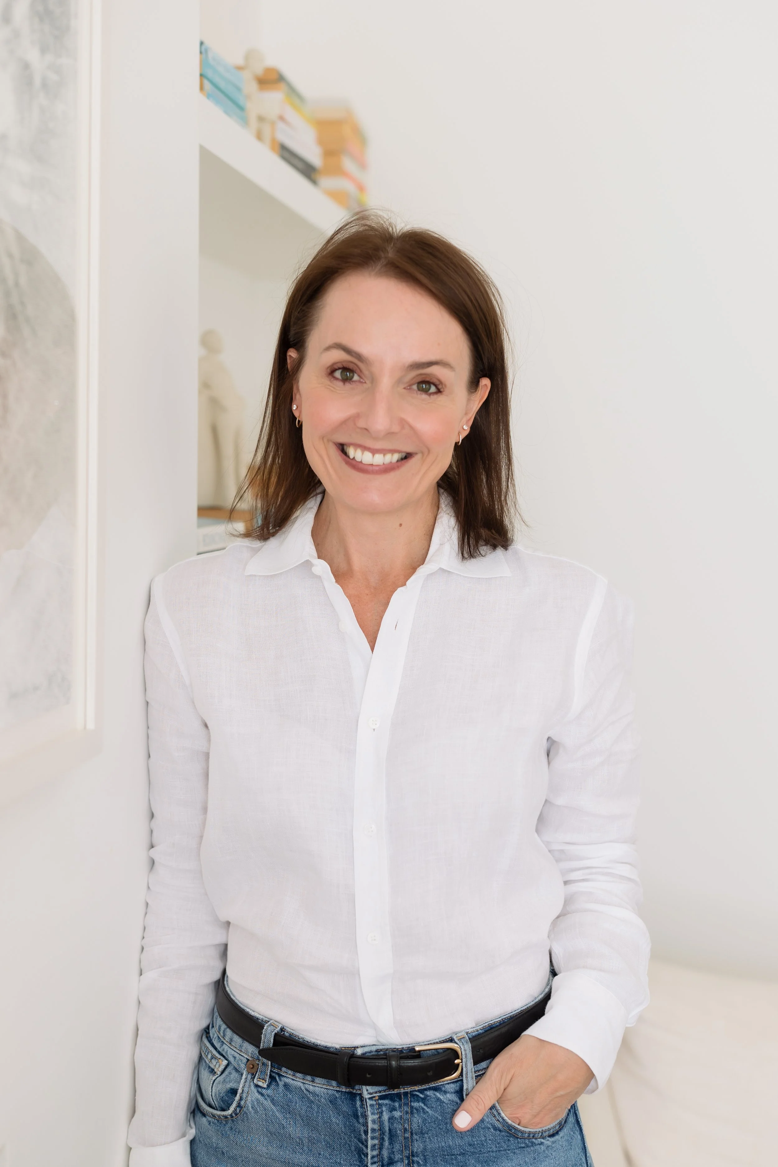 Jeanne Vorster Naidoo, Clinical Psychologist, standing relaxed against a white wall in her consulting room with one hand in her pocket, smiling, in a white linen shirt and jeans, with bookshelves softly blurred behind her.