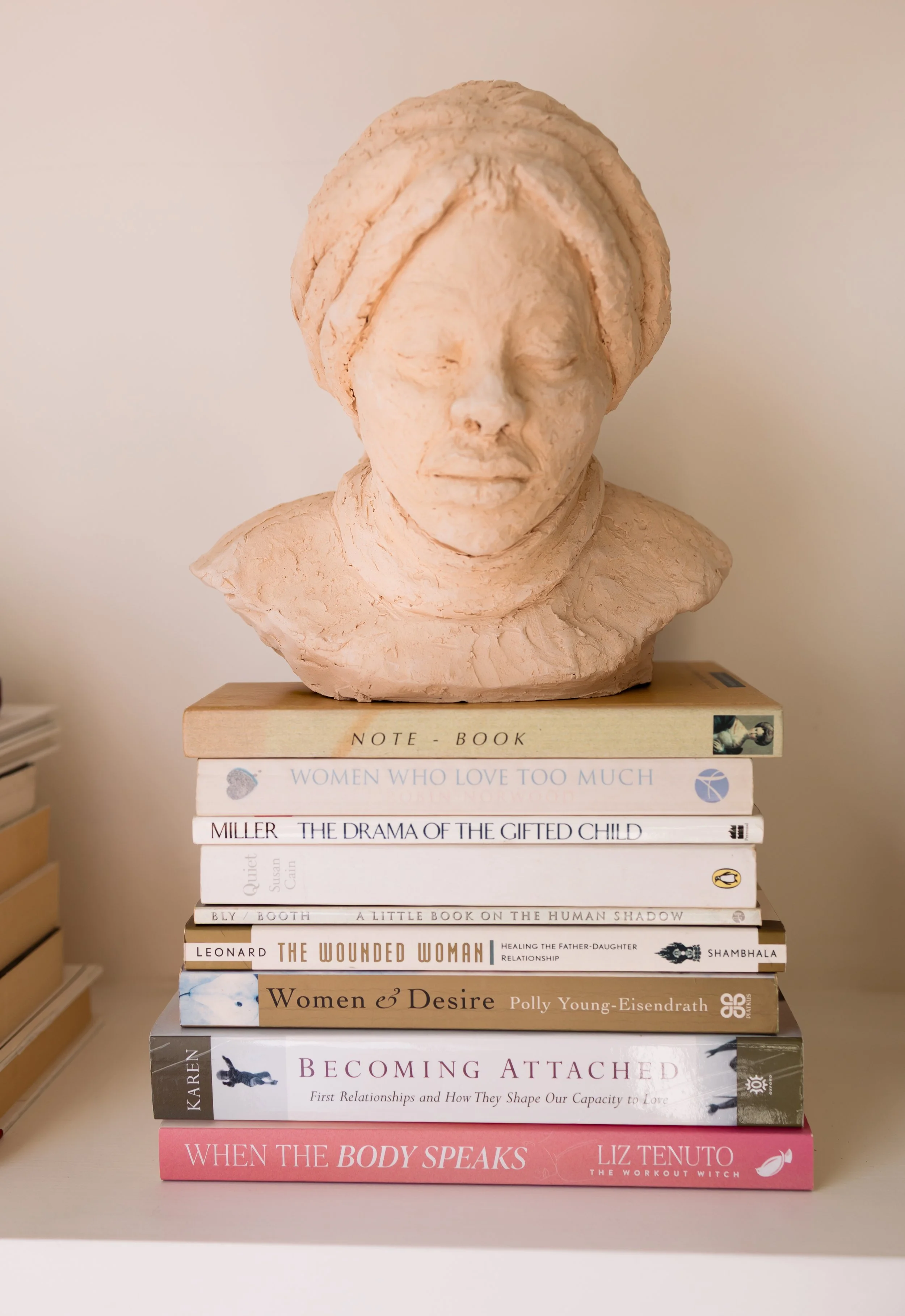 A bust sculpture of a woman wearing a headscarf, placed on top of a stack of books. The books include titles about women, relationships, and personal development.