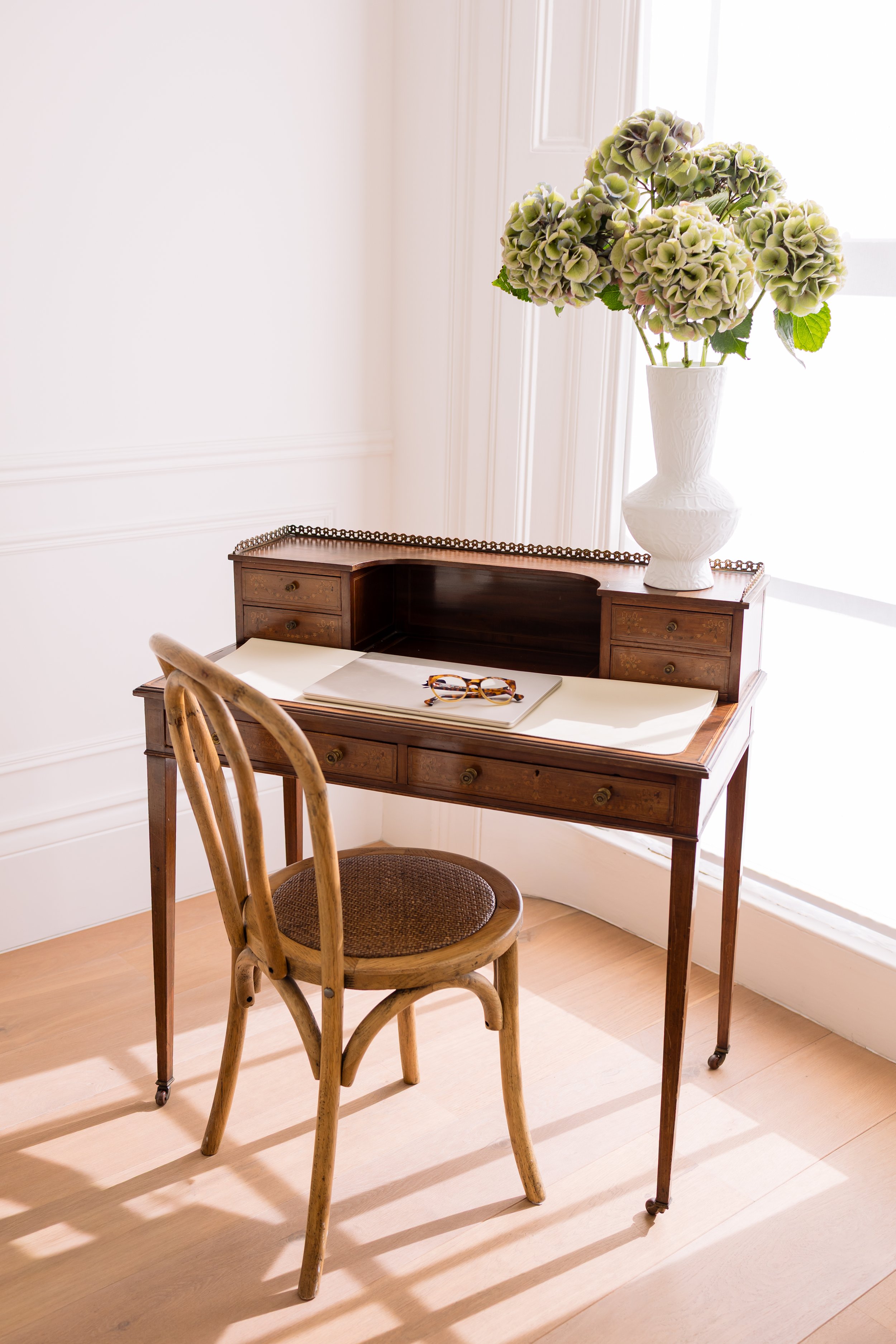 A wooden desk in St John's Wood consulting room with an open book and a pair of glasses, a wooden chair with a woven seat, and a large white vase with green hydrangea flowers in front of a window with white curtains.
