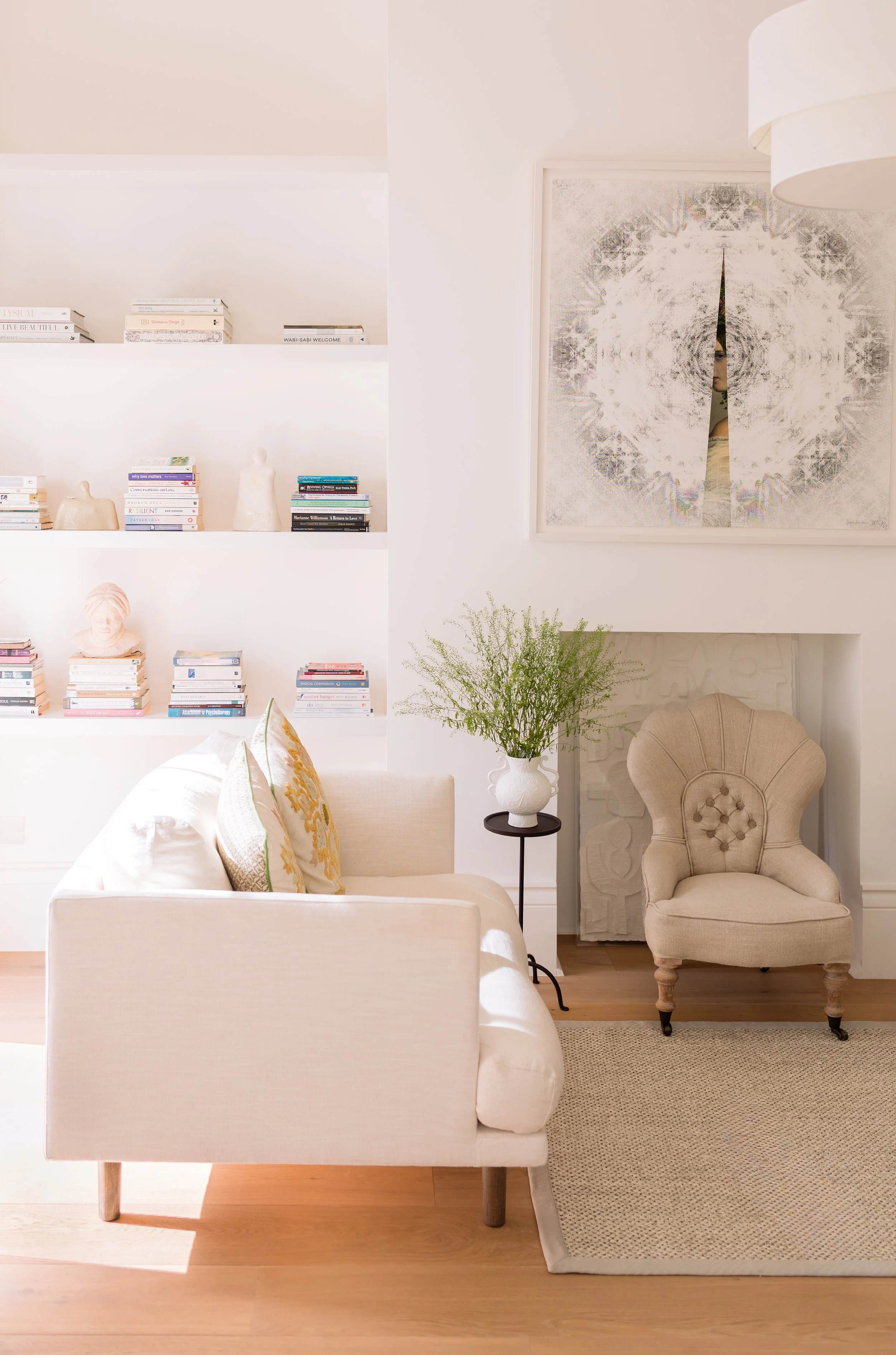 A calm consultation room in St John's Wood with a white sofa, decorative pillows, an armchair, a side table with a white vase and plant, a bookshelf filled with books and sculptures, a large abstract wall.