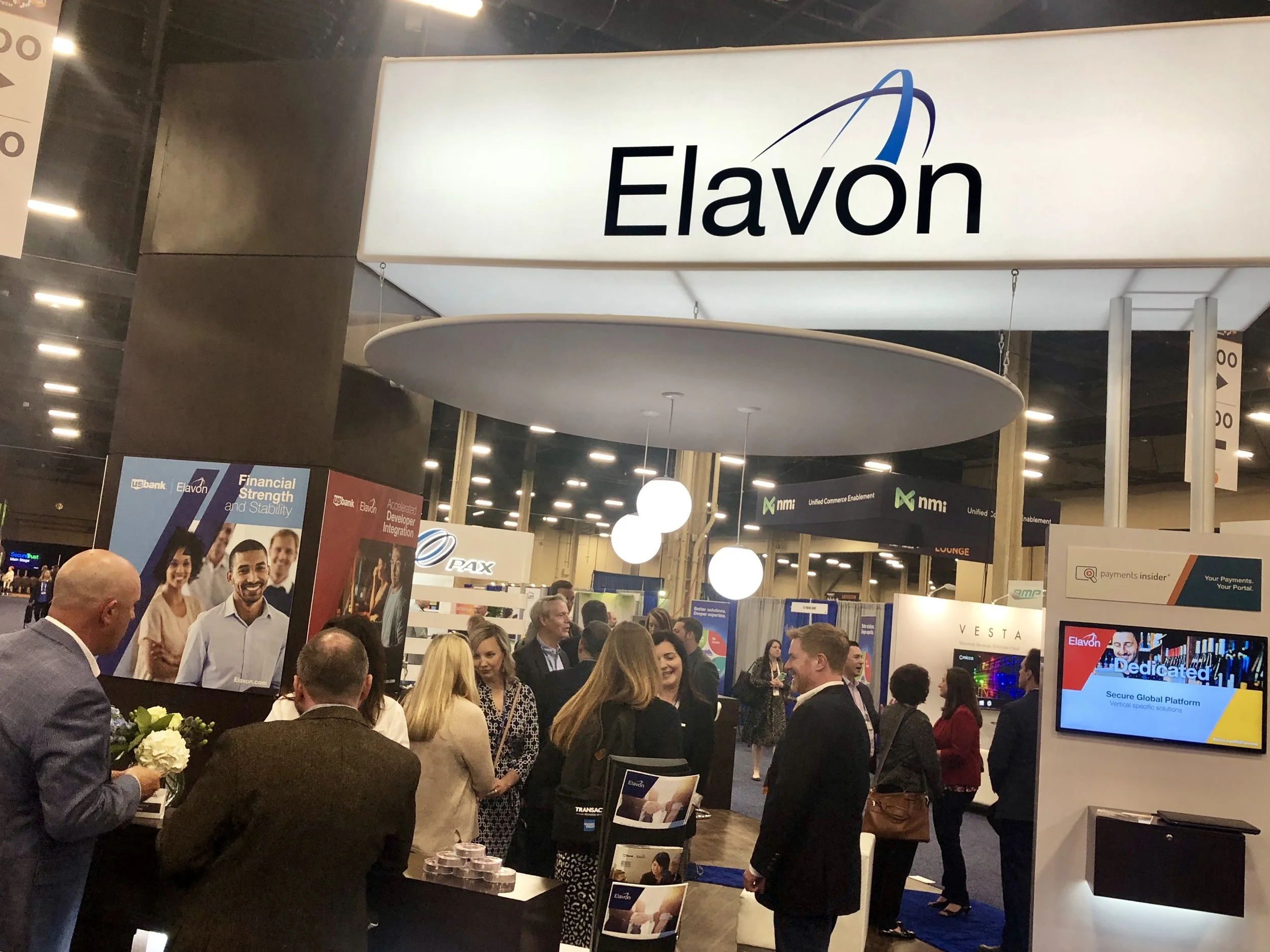 A busy trade show or conference with many attendees gathered around the Elavon booth. The booth has a large white sign with the Elavon logo and name. There are promotional displays, screens, and people engaging in conversations.