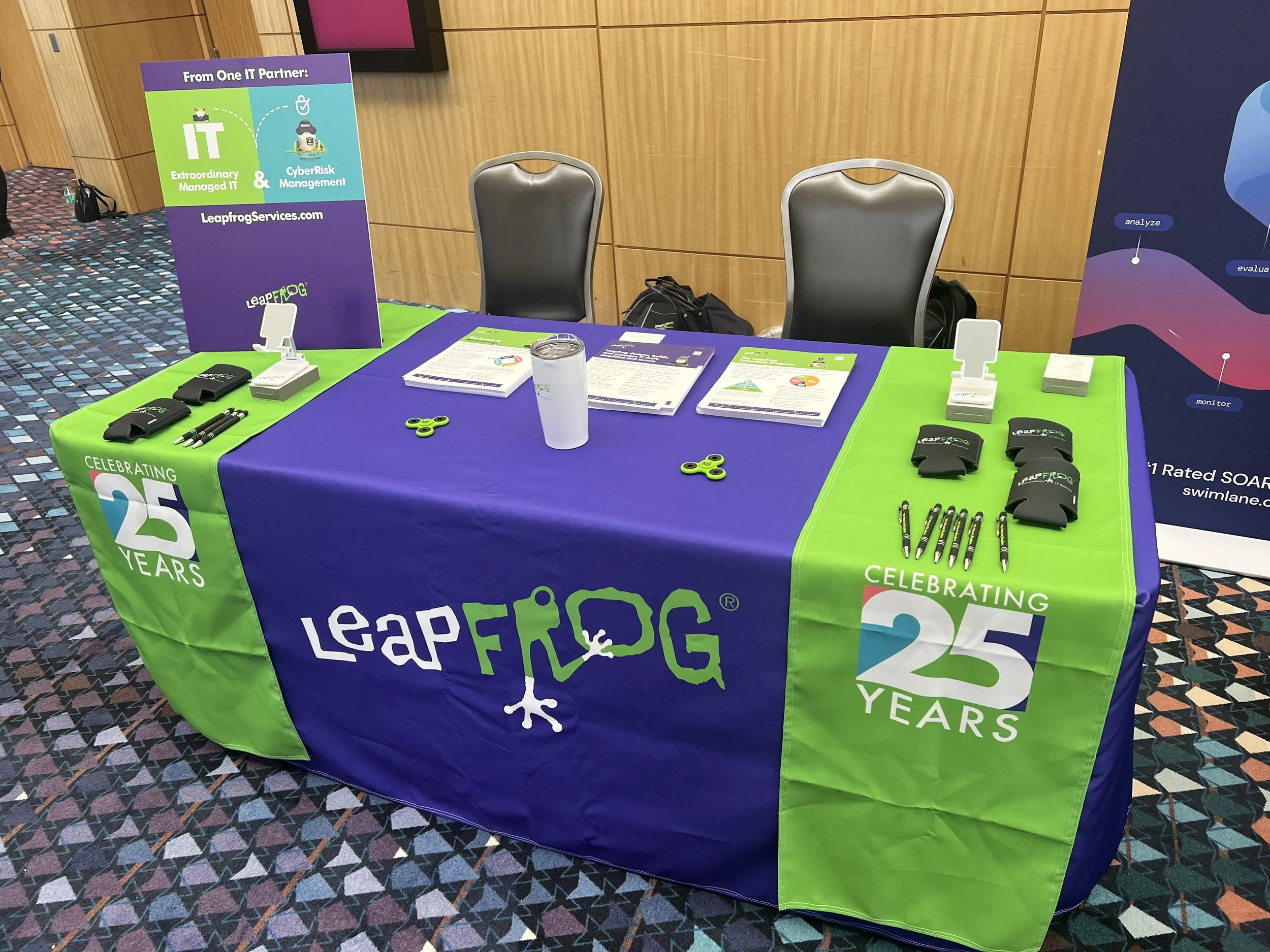 Exhibition booth table with marketing materials for Leapfrog Services, celebrating 25 years, featuring promotional items like pens, fidget spinners, and brochures, set against a wood-paneled wall and a patterned carpet.