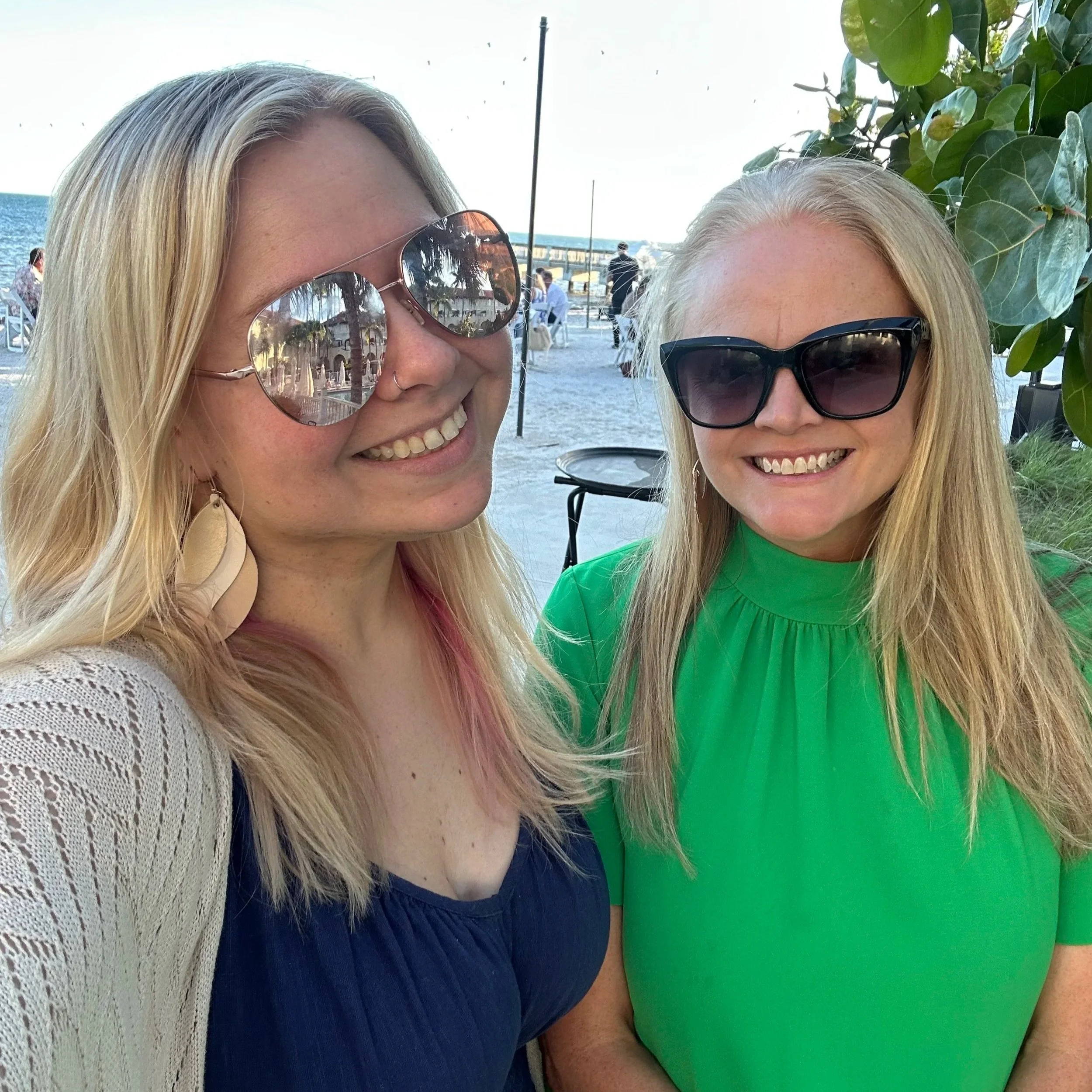 Two women smiling at the camera, wearing sunglasses, outdoors near a body of water, with other people in the background.