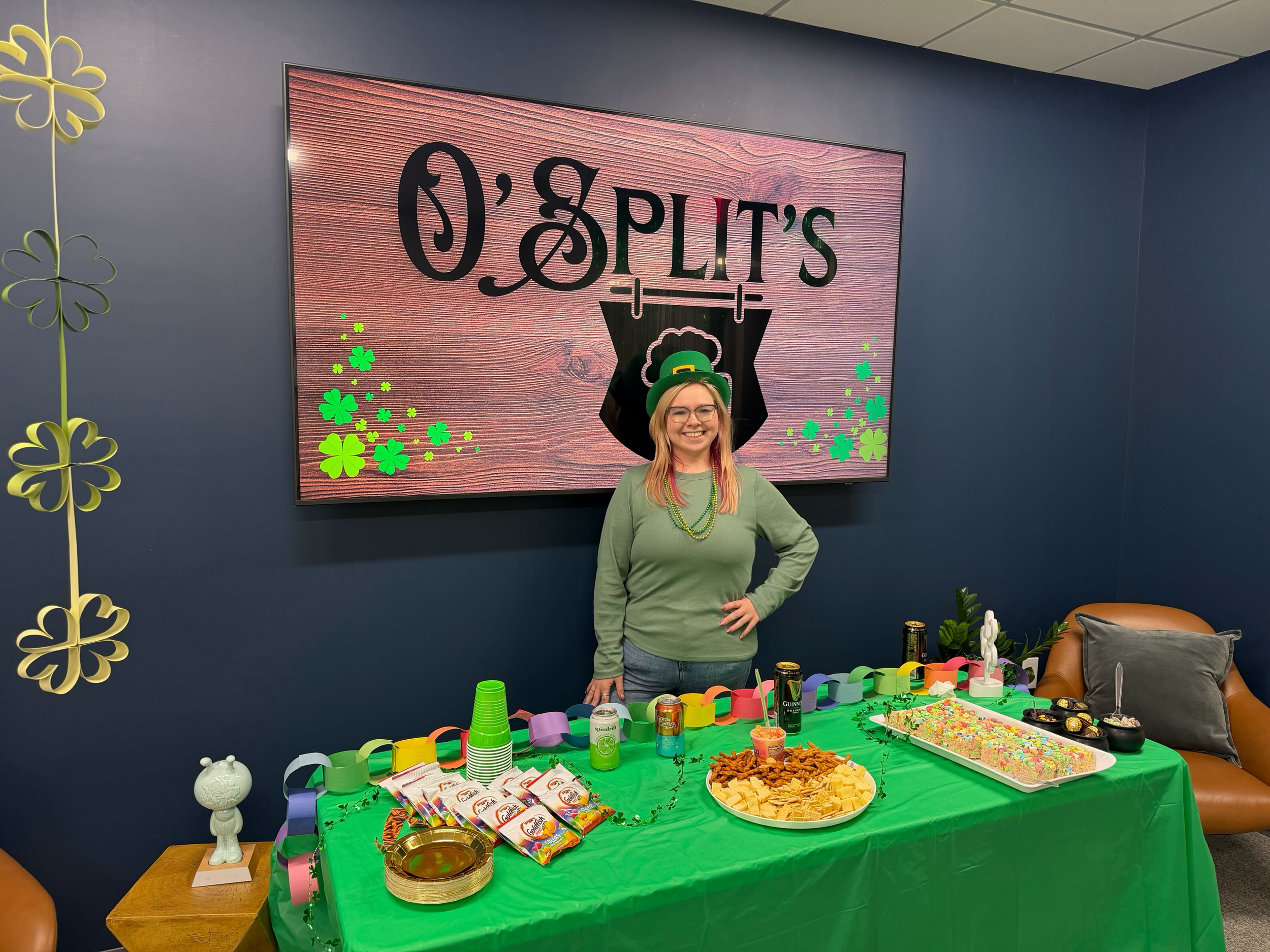 A woman wearing a green hat and green beads stands behind a table decorated with a green tablecloth, set with snacks, candies, and cupcakes for a St. Patrick's Day celebration. The table is decorated with shamrock and rainbow paper chains. Behind her is a decorated wall with shamrocks and a large sign that reads "O'Spirit's" for a St. Patrick's Day party.
