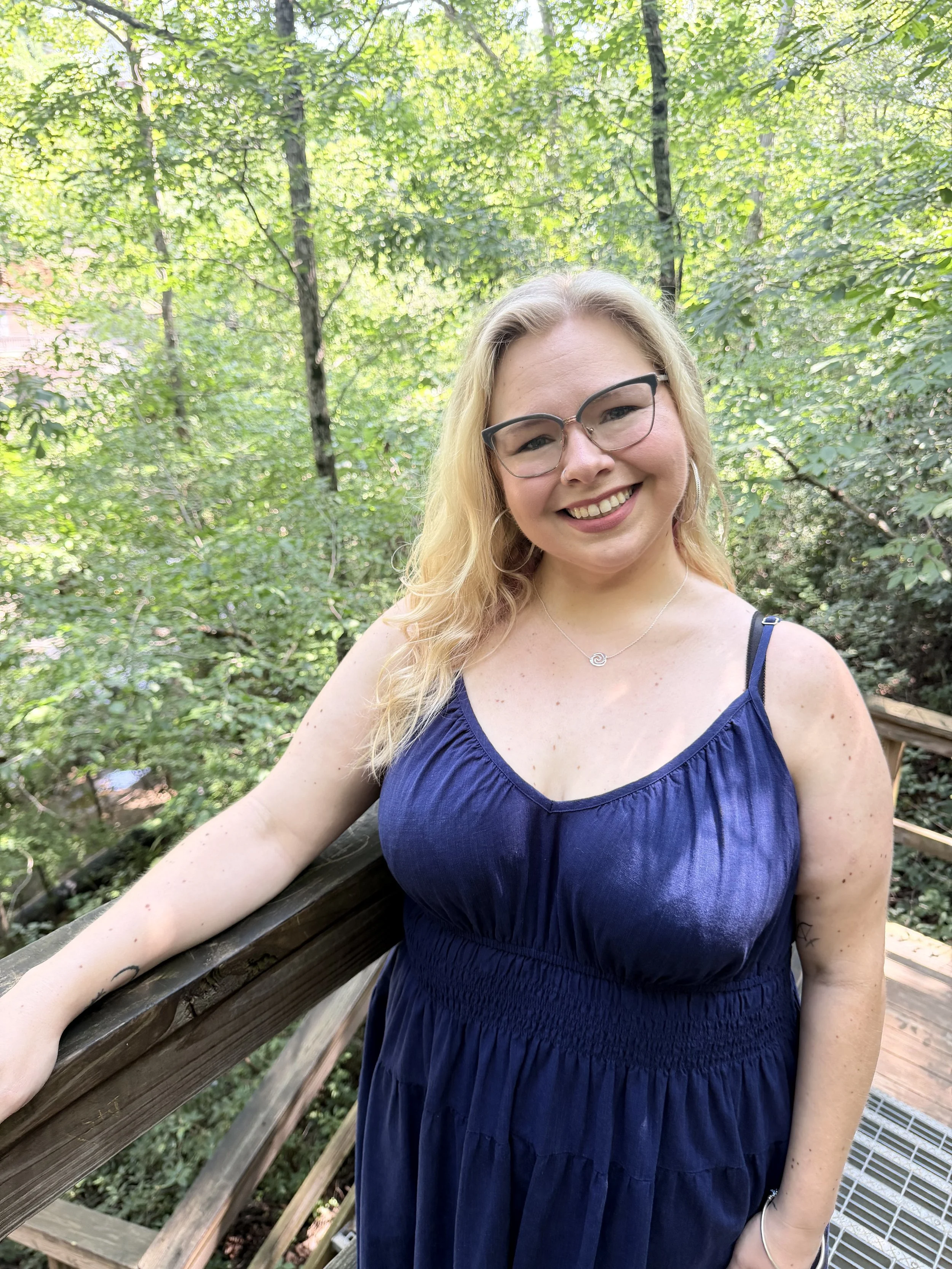 A woman with glasses and blonde hair smiling at the camera on a wooden bridge or railing in a lush green forest.