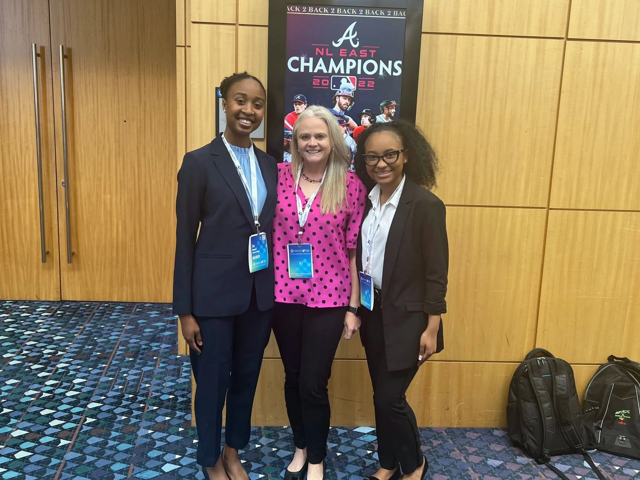 Two women and one woman in a pink polka dot shirt standing together at an event with a poster behind them that reads "NL East Champions 2022" with images of baseball players.