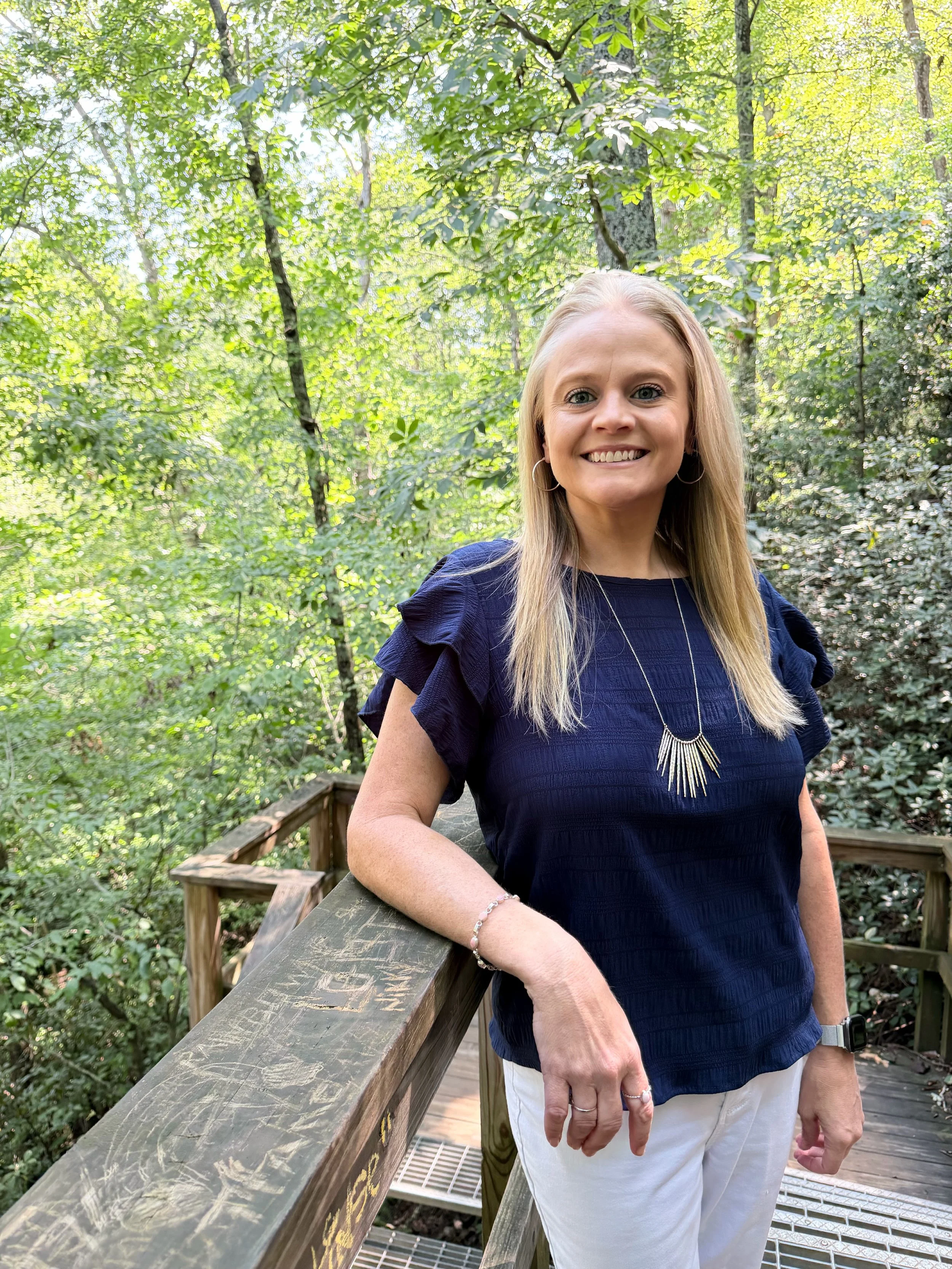 A woman with blonde hair smiling while standing on a wooden bridge in a green forest, wearing a navy blue blouse, white pants, and jewelry.